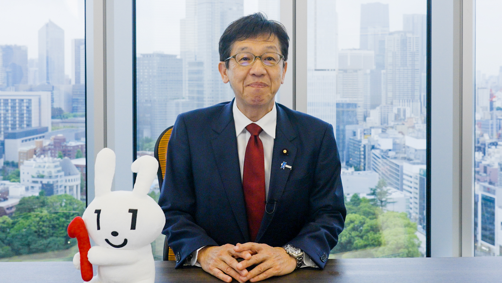 A photo of Minister for Digital Transformation Matumoto sitting on a chair with his hands folded on a desk decorated with a stuffed Maina-chan doll.