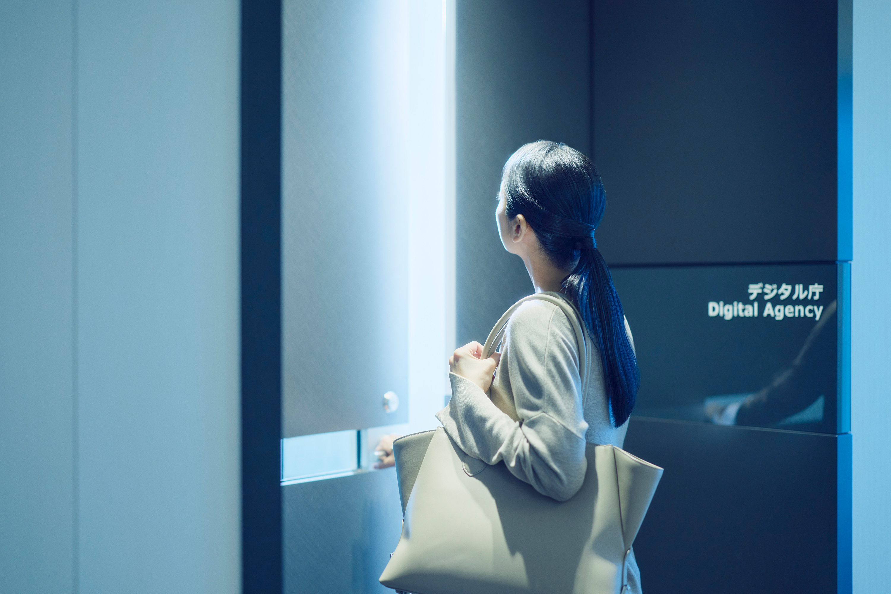 デジタル庁のオフィス入口前に立つ女性の後ろ姿の写真。女性は入口に向かって立っており、右手をドアノブにかけ、これから入室する様子。長い黒髪をポニーテールにまとめ、ベージュのニットを着用し、白いトートバッグを肩にかけている。右手前方には「デジタル庁 Digital Agency」と日英併記された黒い壁が見える。オフィス内部からは光が差している。