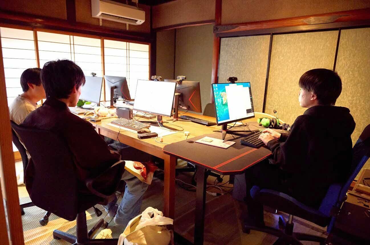 障子や木の梁・長押が特徴的な和室のしつらえの、島根県デジタル拠点サービス提供者認定拠点の中に、ゲーミングデスクと複数台のモニターが設置されている。黒い服装の若い男性3人がゲーミングチェアに座り、横並びになり、アプリ開発やeスポーツに取り組んでいる。デスク上にはキーボード、ウェブカメラ、緑色のゲーミングヘッドセット、ゲームパッドなどがある。