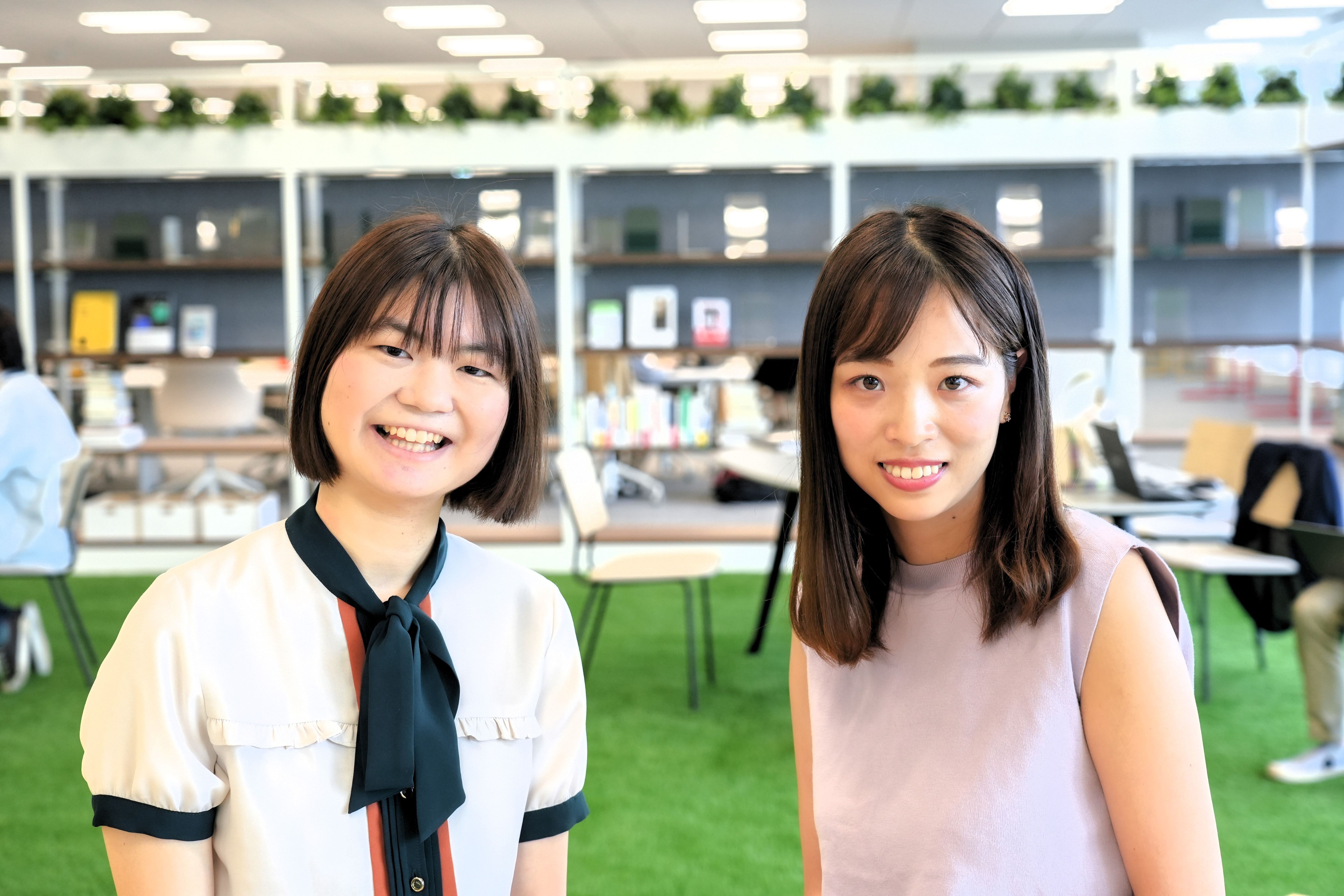 Two women are smiling, facing the front. They are shown from the chest up. The woman on the left is wearing a whitish one piece dress. The woman on the right is wearing a pink sleeveless one piece dress. The background is an office artificial lawn or a bookshelf. 