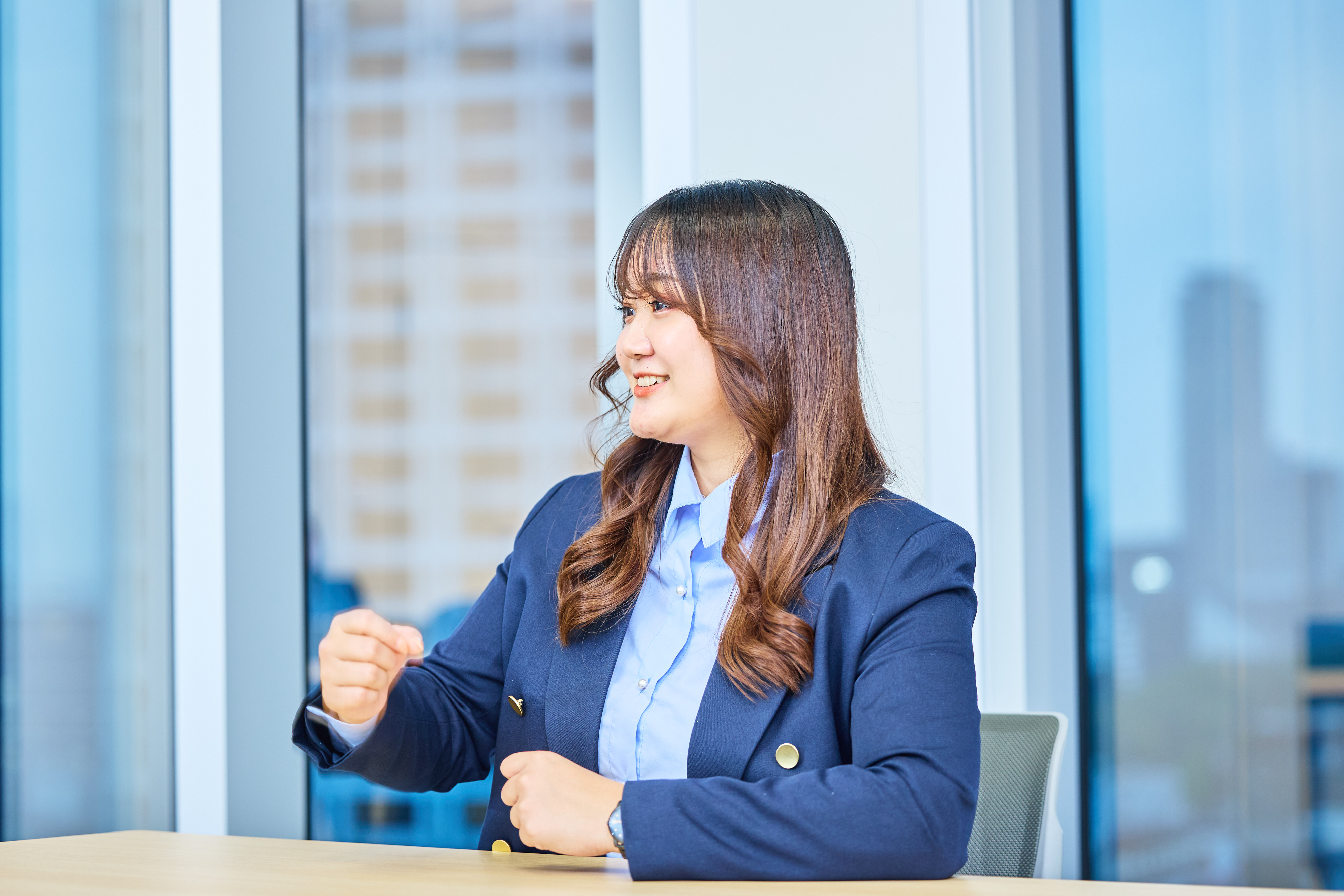 A woman gestures, smiling, talking to her left. She is wearing a navy jacket and a blue shirt. The place is a conference room. She sits at a wooden table. In the background, a window glass shows Bill. 