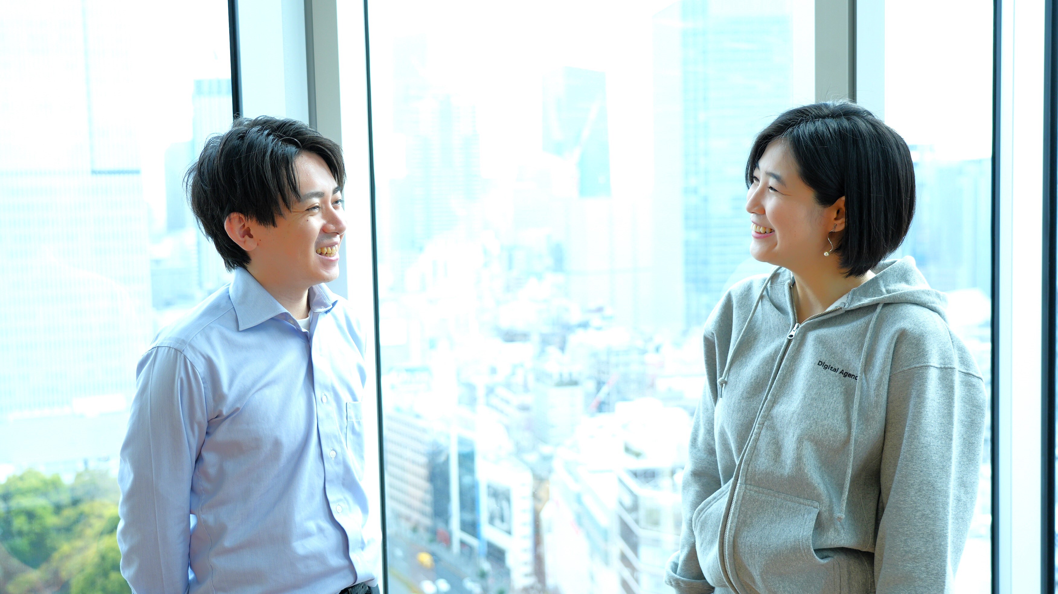 A man and a woman face each other, smiling. They are shown from the waist up. To the left is a man, wearing a light blue shirt. To the right is a woman, wearing a gray hoodie. The background is a windowpane showing Bill. 