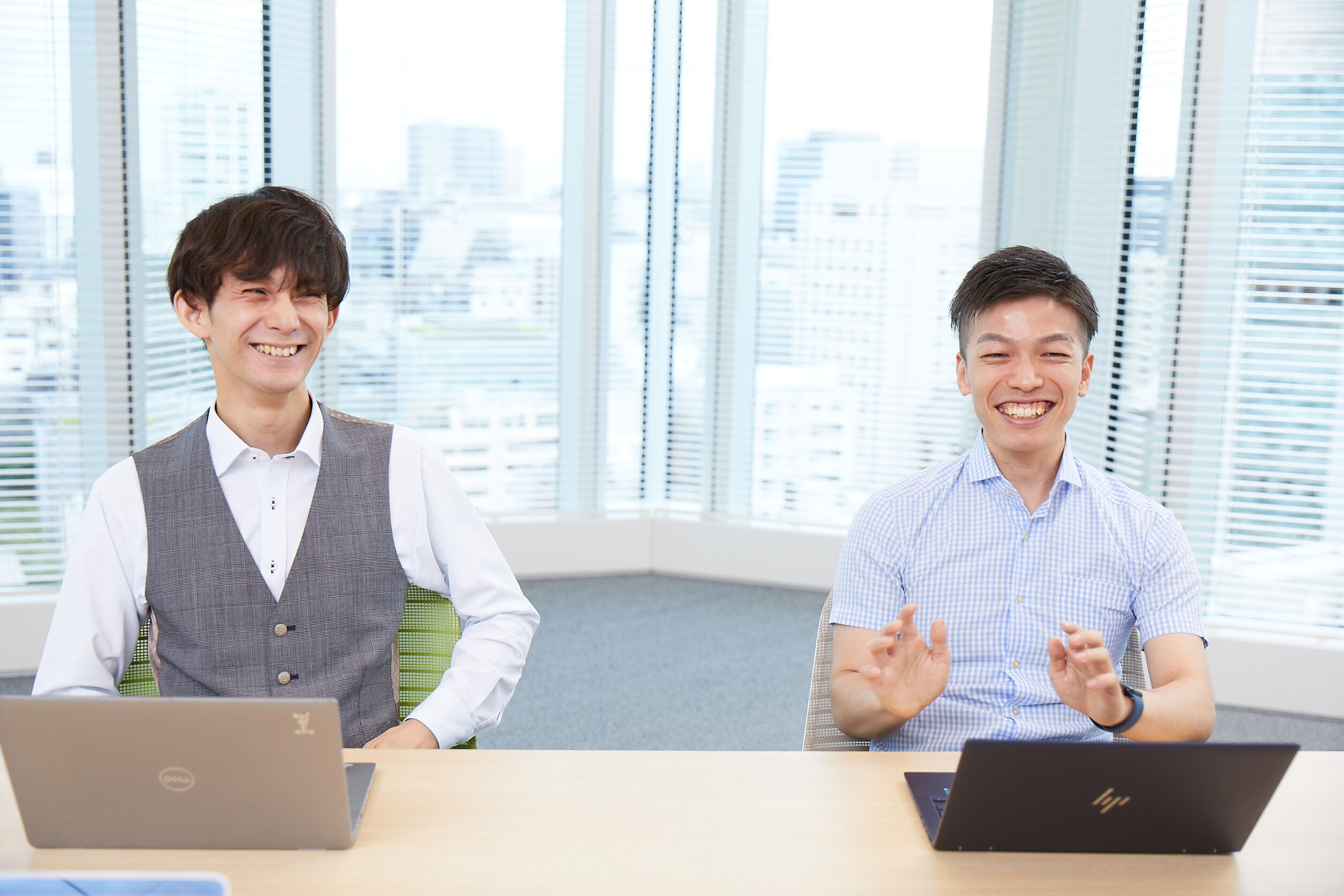 Two men face each other, smiling. They are sitting at desks, each with a laptop in front of them. The man on the left is wearing a white shirt with a gray vest. The man on the right is wearing a light blue short-sleeved striped shirt. The background is a windowpane showing Bill. 