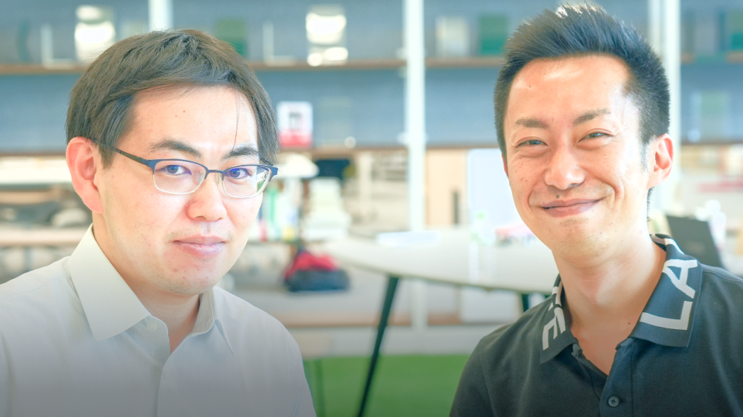 Two men standing side-by-side are pictured from the neck up. The man on the left is wearing a white shirt and glasses. The man on the right is wearing a black polo shirt and is smiling. In the background is an office bookshelf. 