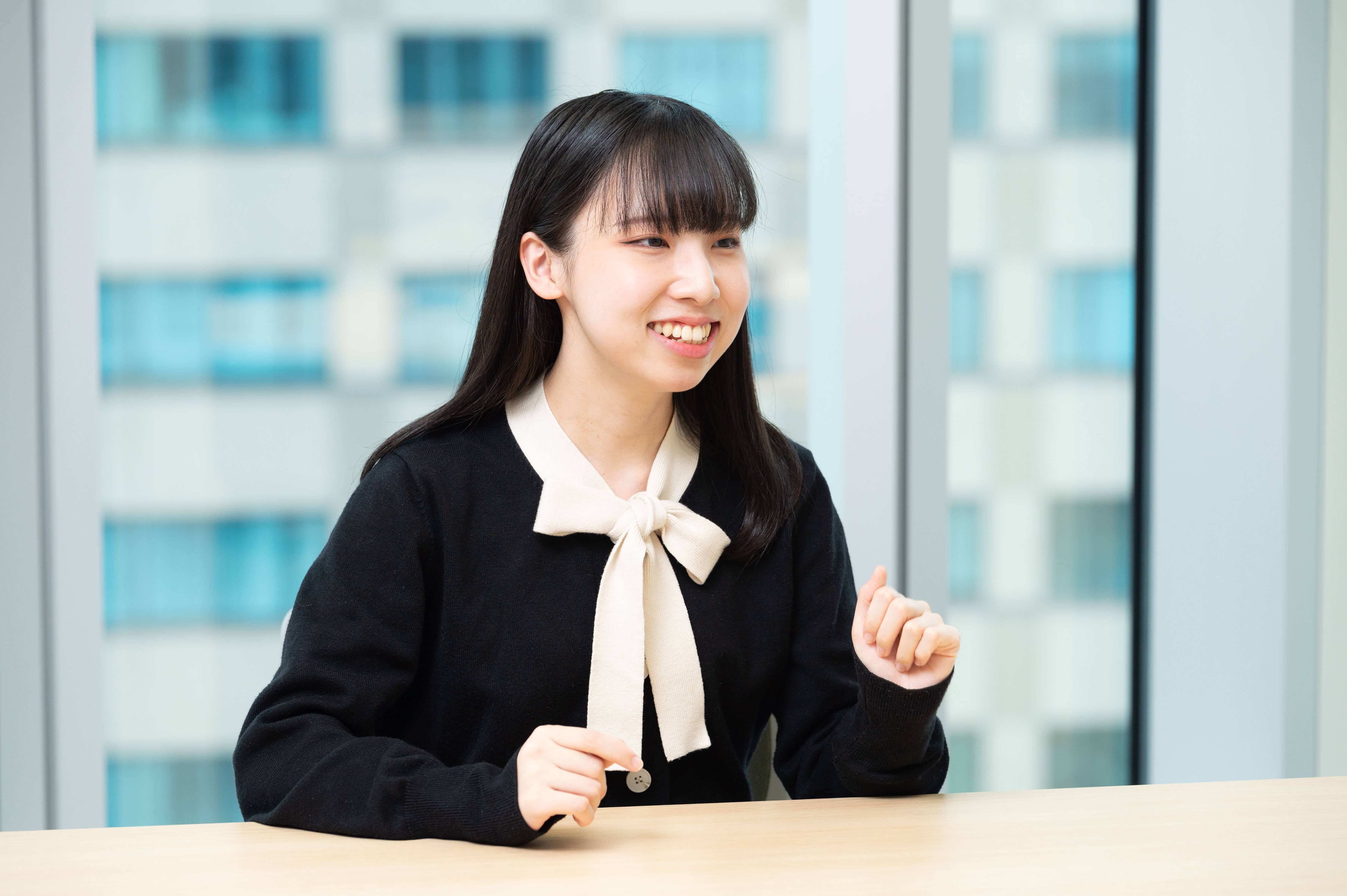 A woman gestures, smiling and talking to her right. She wears a black dress with a white ribbon around her neck. The place is a conference room. She sits at a wooden table. In the background, a window glass shows Bill. 