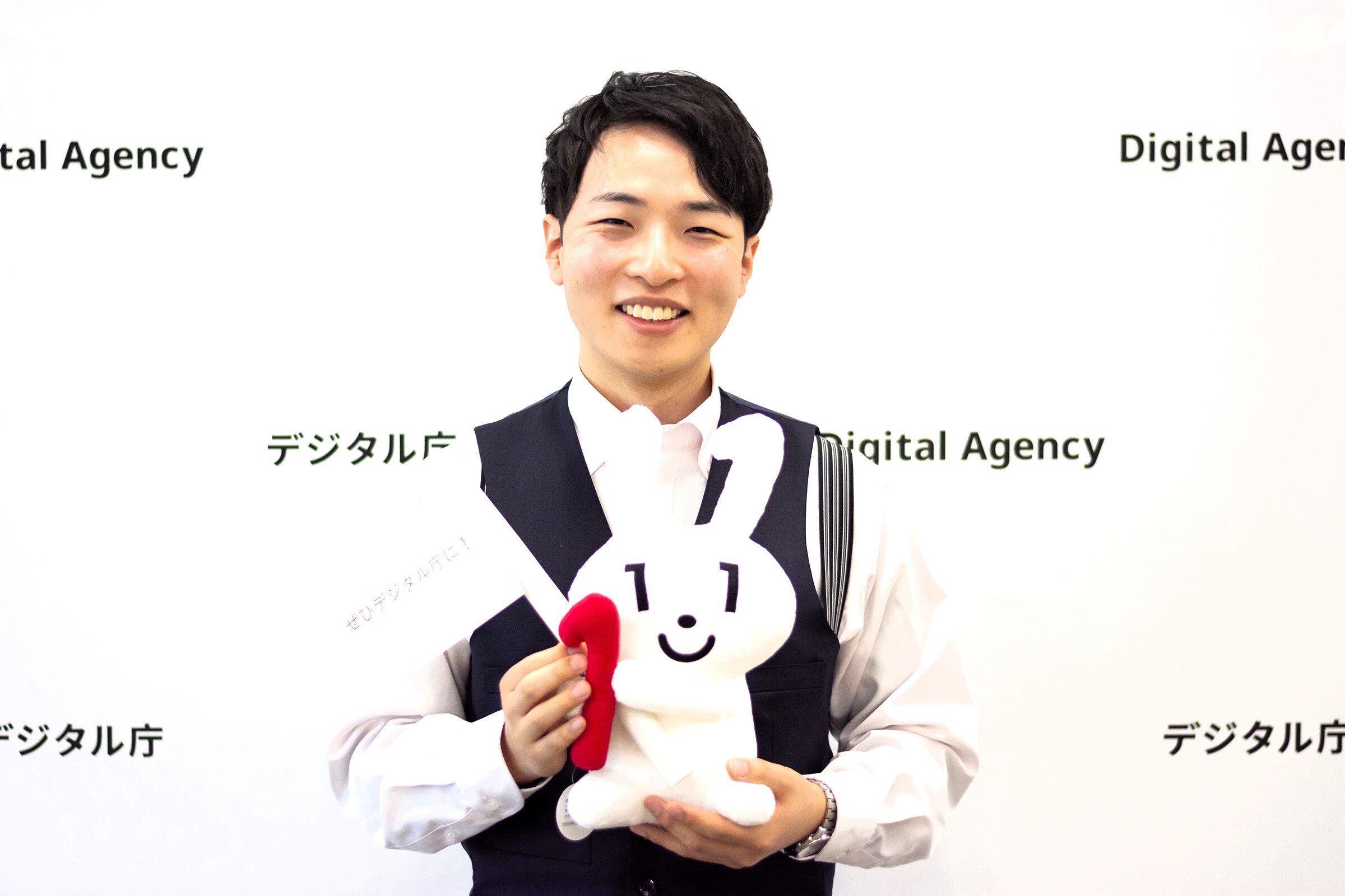 A smiling man faces the front, smiling as he carries his Maina-chan. He wears a white shirt and a black vest. The location is the Digital Agency office, with the words 