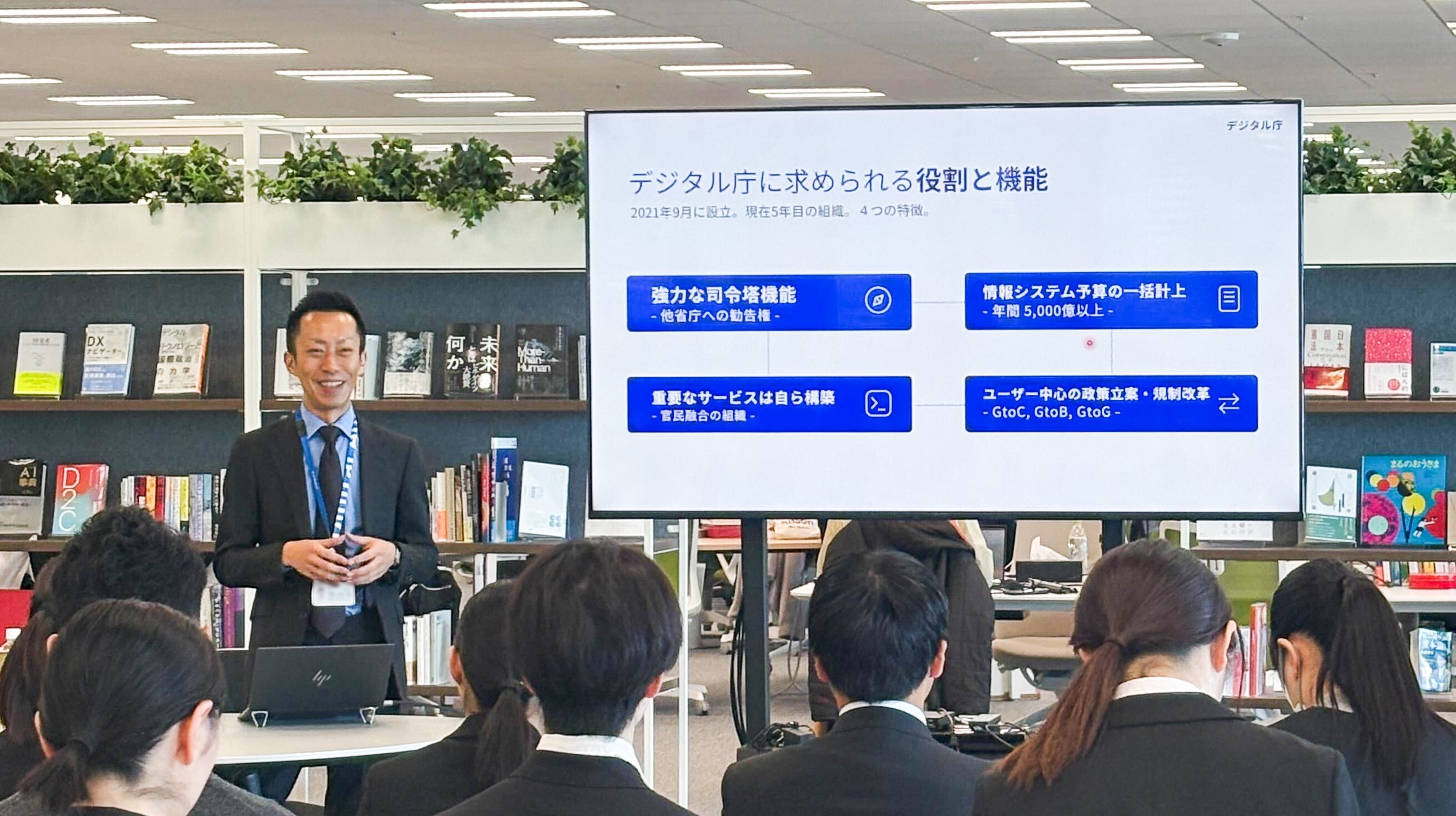 A photo of a briefing session&comma; with a large monitor in front and a person standing next to it&comma; smiling and talking to the audience&period; 