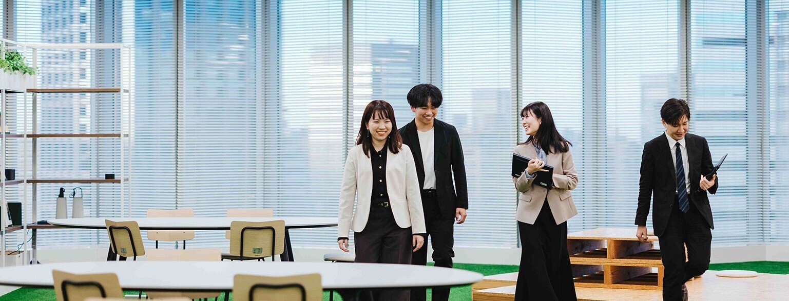 Four Digital Agency officials are pictured walking in the wide space of the office. Large windows in the background offer views of the city, and round tables and chairs are arranged around the perimeter. The floor is a green carpet, and the ceiling has multiple lights