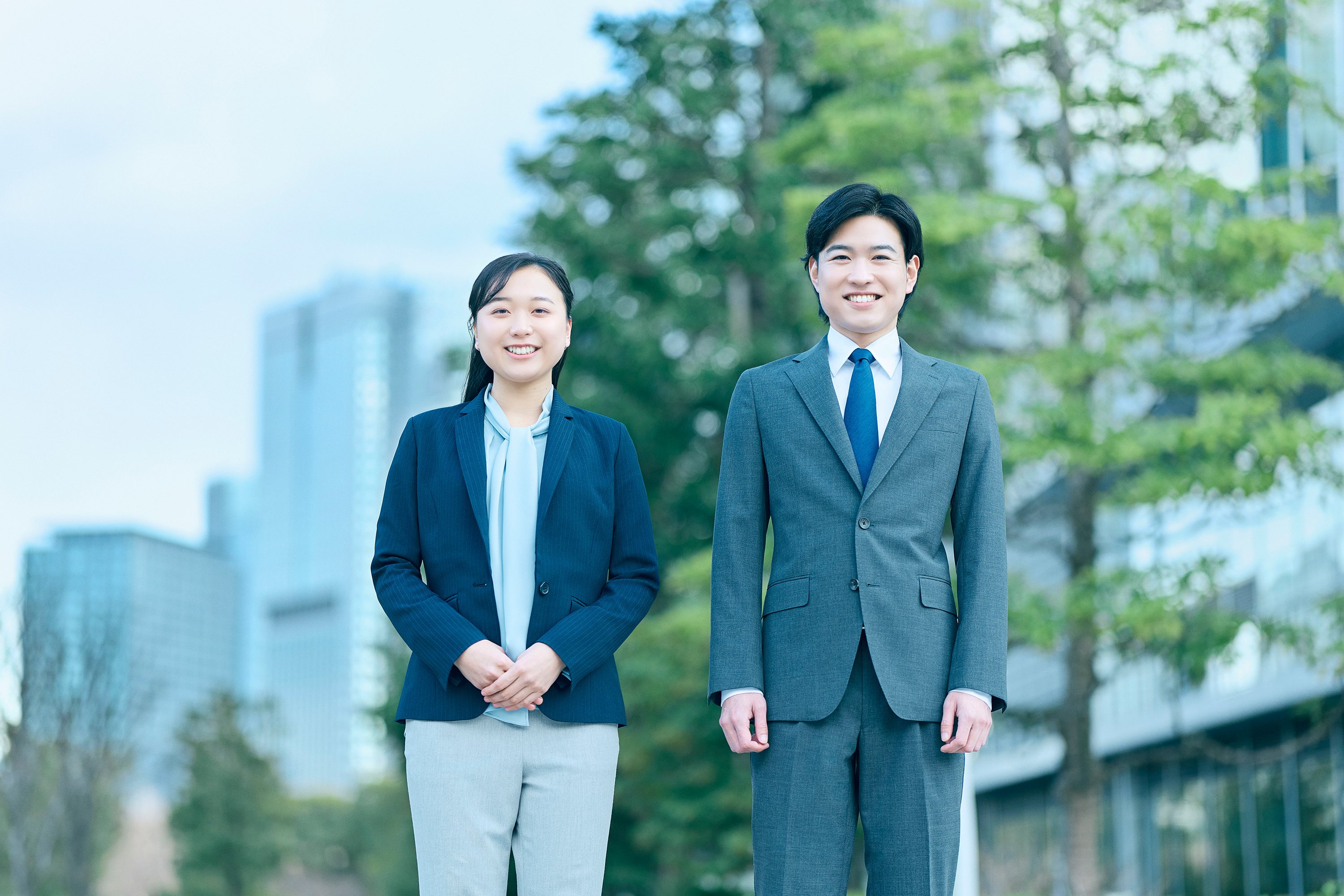 屋外のビジネス街で、ビジネススーツを着た男女2名が並んで立っている写真。左側の女性は紺色のジャケットに水色のブラウス、ライトグレーのパンツを着用し、両手を前で組んで笑顔を見せている。右側の男性は青いネクタイにグレーのスーツを着用し、両腕を体の横に下ろして笑顔で立っている。背景には緑豊かな街路樹とオフィスビルが見える。
