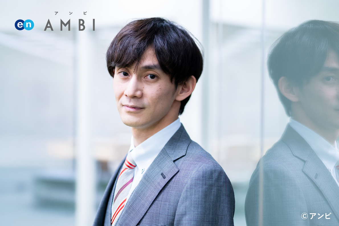 男性職員がガラス窓の前に立つ写真。左上に「en AMBI」のロゴがあり、右下に著作権表示「©アンビ」が記載されている。