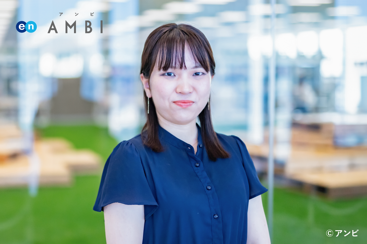 女性職員が室内のオフィス内で立っている写真。左上に「en AMBI」のロゴがあり、職場やオフィス環境を示している。