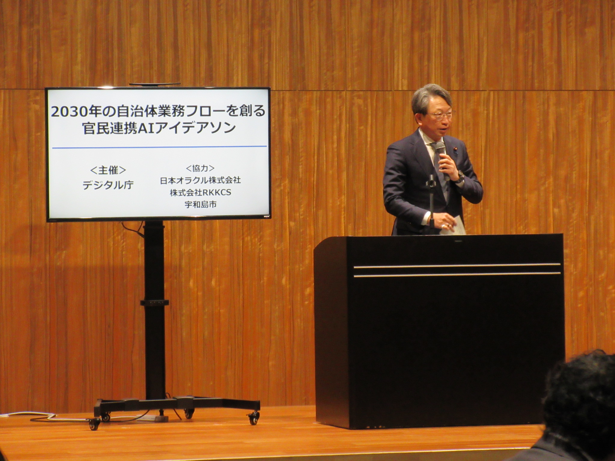 Mr. Taira, Minister for Digital Transformation, is shown on the right, and a monitor is shown on the left, showing Mr. Taira, Minister for Digital Transformation, talking. 