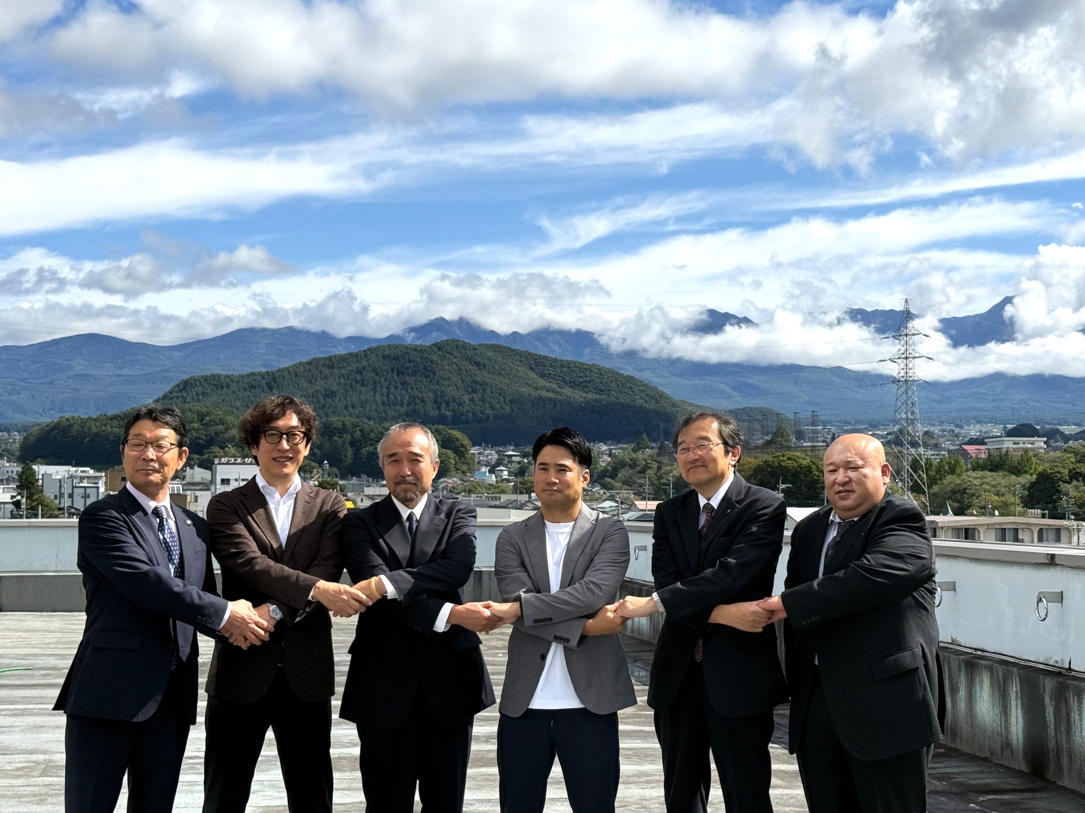 Outside the Chino City Hall in Nagano prefecture, with mountains and a blue sky in the background, from left to right, Mayor Atsushi Imai of Chino-shi, Coordinator for Planning of Toriyama, Group of Service for Citizens, Digital Agency, Takeki Yonekawa, representative of Yatsugatake Tourist Association and Kuroyuri Hutte, Daisuke Hokari, representative of Northern Alps Lodge Friendship Association and Mr. Yarigatake Sanso Group, Akira Kumagai, Regional Revitalization Policy Director of Chino-shi, and Iwao Morozumi, vice president of Yatsugatake Tourist Association and representative of Karasawa Mineral Hot Spring, cross their arms and shake hands next to each other.