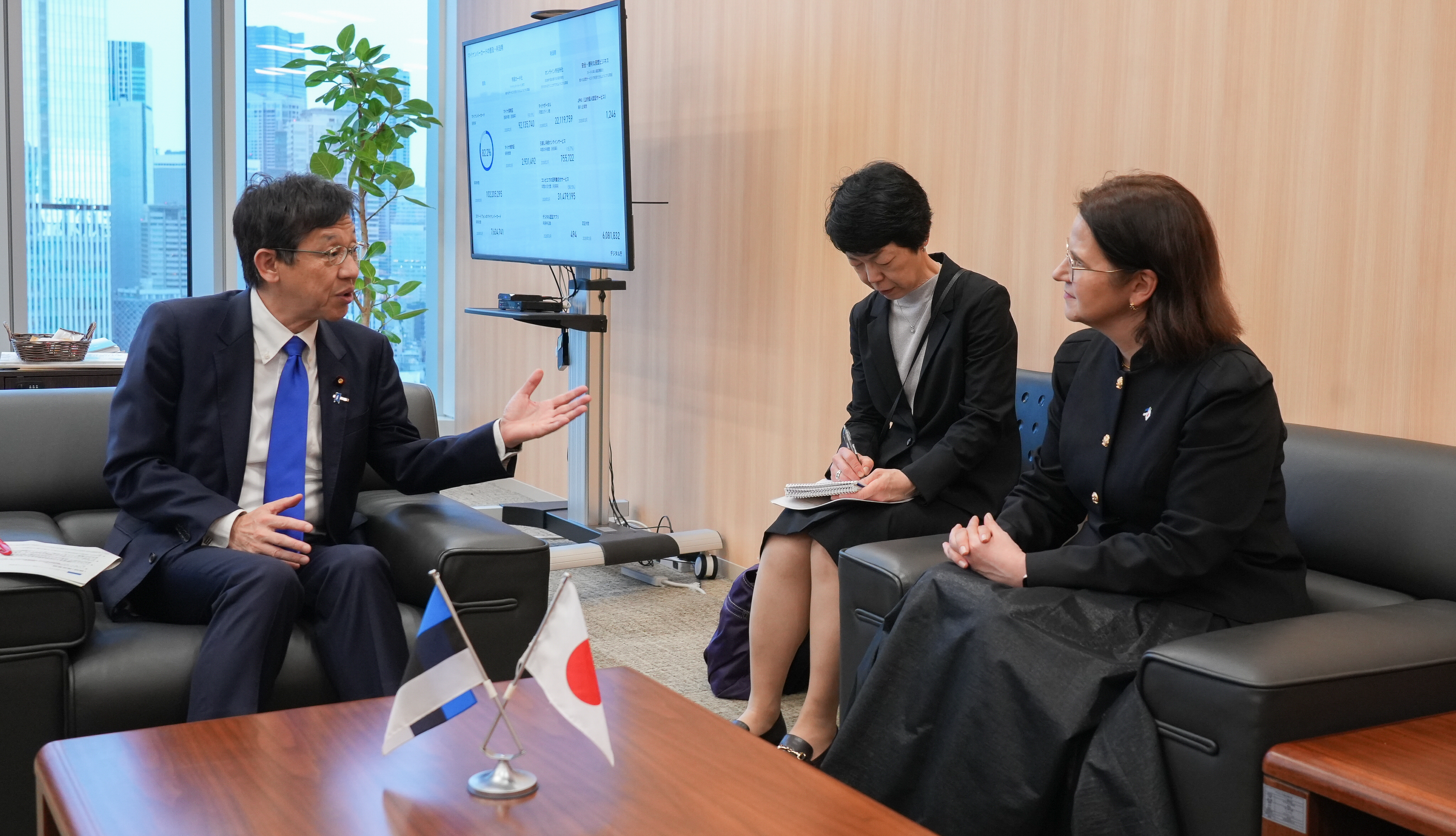 Minister for Digital Transformation Matsumoto sits on the left, wearing a dark navy suit, white dress shirt, and a bright blue tie, gesturing with his hand as he speaks. Opposite him on the right, Ms. Liisa-Ly Pakosta, Minister of Justice and Digital Affairs of the Republic of Estonia, wears a black jacket with gold buttons and a long dark skirt, listening attentively with her hands clasped. Between them, a women interpreter dressed in a black suit with a white blouse looks down while writing notes in a notebook. On the table in front of them are two small desk flags, Japan and Estonia, indicating a bilateral meeting. Behind them, a large window reveals a city skyline, a green potted plant stands nearby, and a monitor on a stand displays charts or data, reinforcing the formal, professional setting of the meeting.