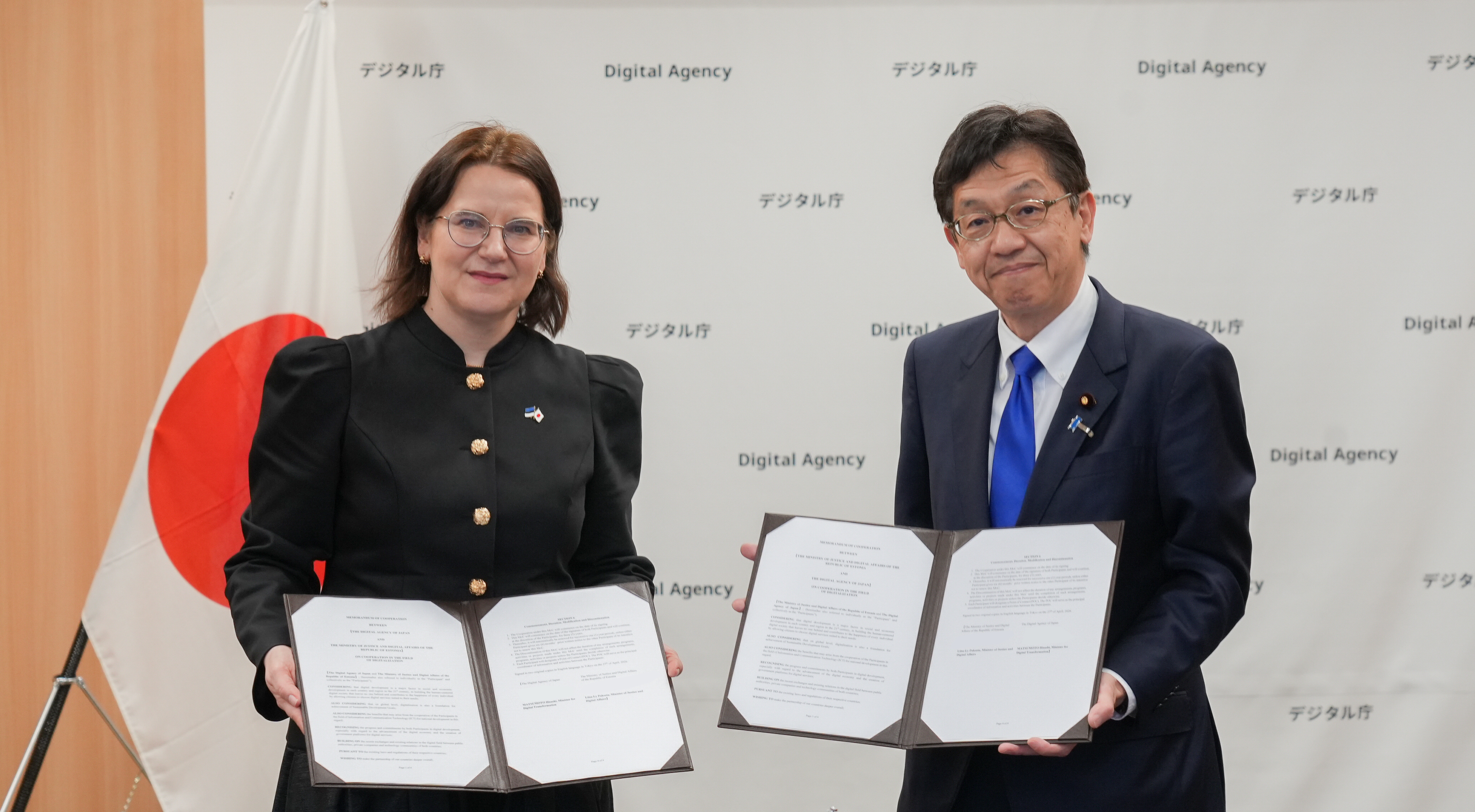 Two ministers stand side by side in a formal setting, each holding open a signed memorandum of cooperation (MoC) folder toward the camera. On the left, Ms. Liisa-Ly Pakosta wears a black long-sleeved dress with gold buttons and glasses. On the right, Minister for Digital Transformation Matsumoto wears a dark suit, white shirt, and bright blue tie. Both faces forward with slight smiles. Behind them is a backdrop reading “Digital Agency,” along with a large Japanese flag.