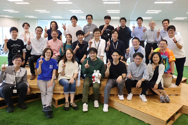 A photo of 26 smiling people standing in three rows with their index fingers raised to represent one team