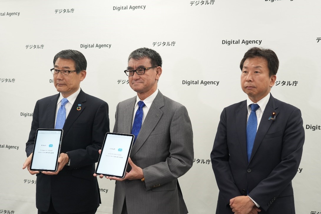 From the left, three people, Chairman Nagamatsu, Minister for Digital Transformation Kono and Senior Vice Minister Okushi Digital, are lined up in a waist shot. Against the background of a bag studded with the Digital Agency logo, Chairman Nagamatsu and Minister for Digital Transformation Kono hold up tablets displaying the agreement.