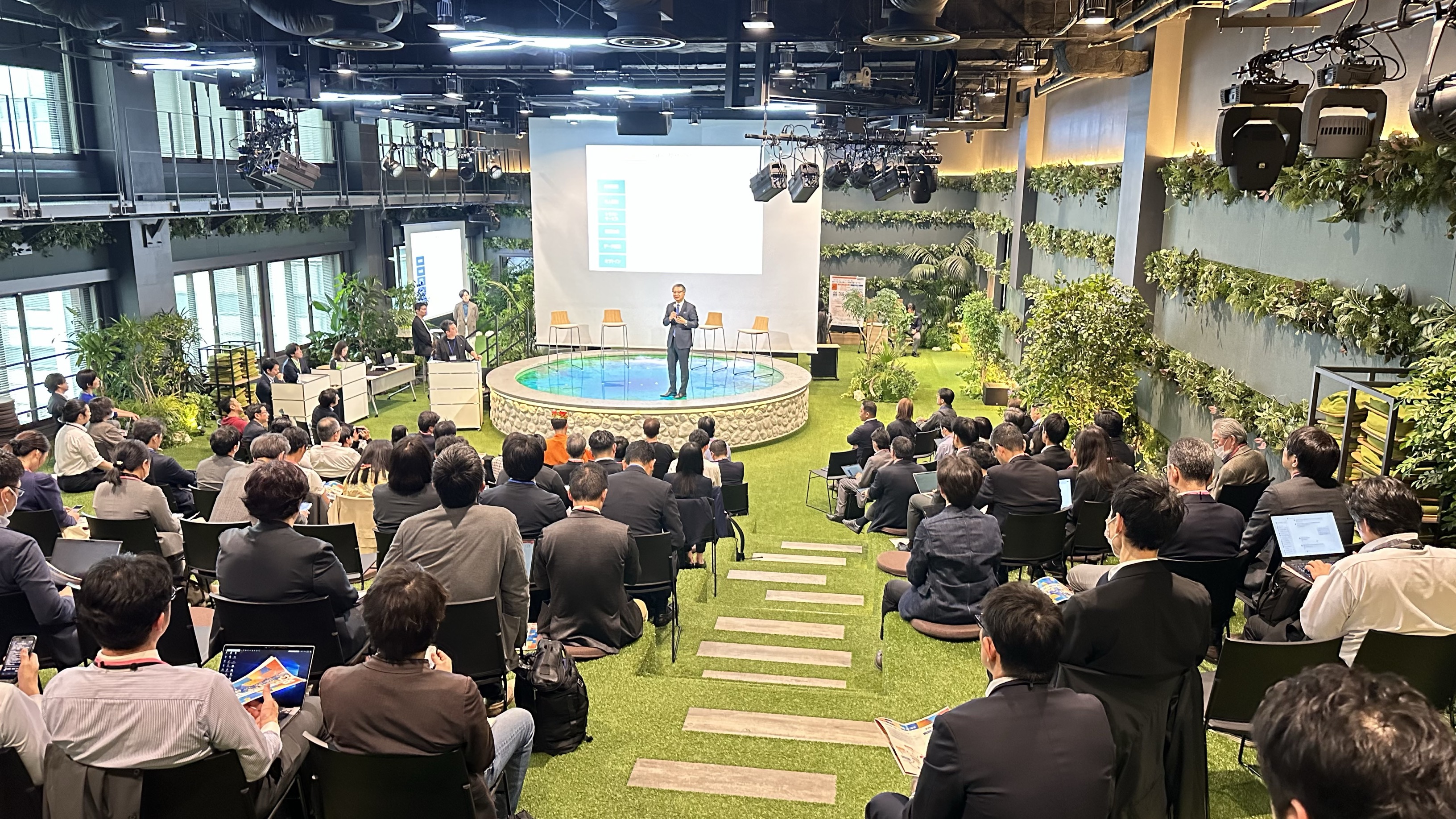 A scene of the entire hall. The presenter is standing, facing the audience and talking. Behind the presenter, there is a projector that projects materials. The floor is stepped and covered with grass. Many spectators are participating, sitting on steps and chairs made of grass and listening to the story.