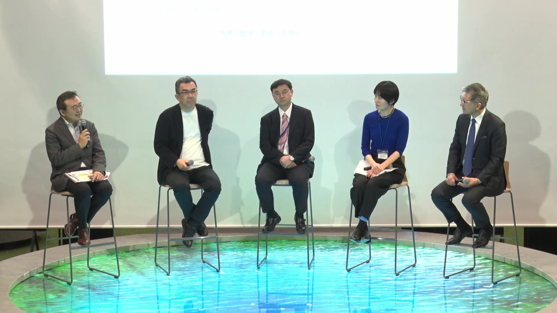 Five people are sitting side by side facing the audience. To the left, Director-General Digital Agency, the moderator, is holding a microphone and talking. Behind the panelists, there is a projector that projects materials. 