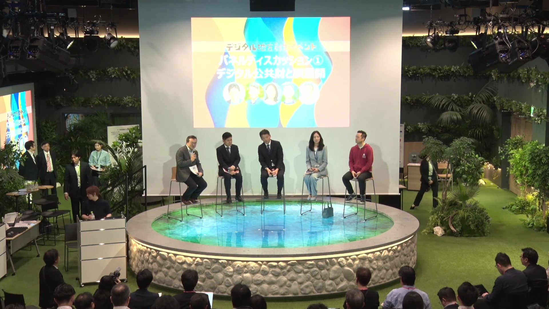 Five panelists are sitting side by side facing the audience, and Director-General Murakami is talking on the microphone. There is a projector behind the panelists to project materials. The venue has a lawn floor and various green plants on the walls and floor. 