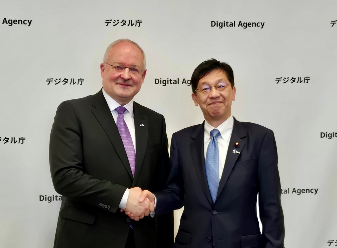 Mr. Andres UNGA, Ambassador of the Republic of Estonia to Japan (left) and Minister for Digital Transformation MATSUMOTO (right), smiling and shaking hands in front of a backdrop featuring a repeating Digital Agency logo.