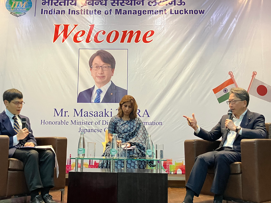 Photo of Minister Taira (right) delivering speech at the Indian Institute of Management (IIM) with a welcome message displayed on the back panel.