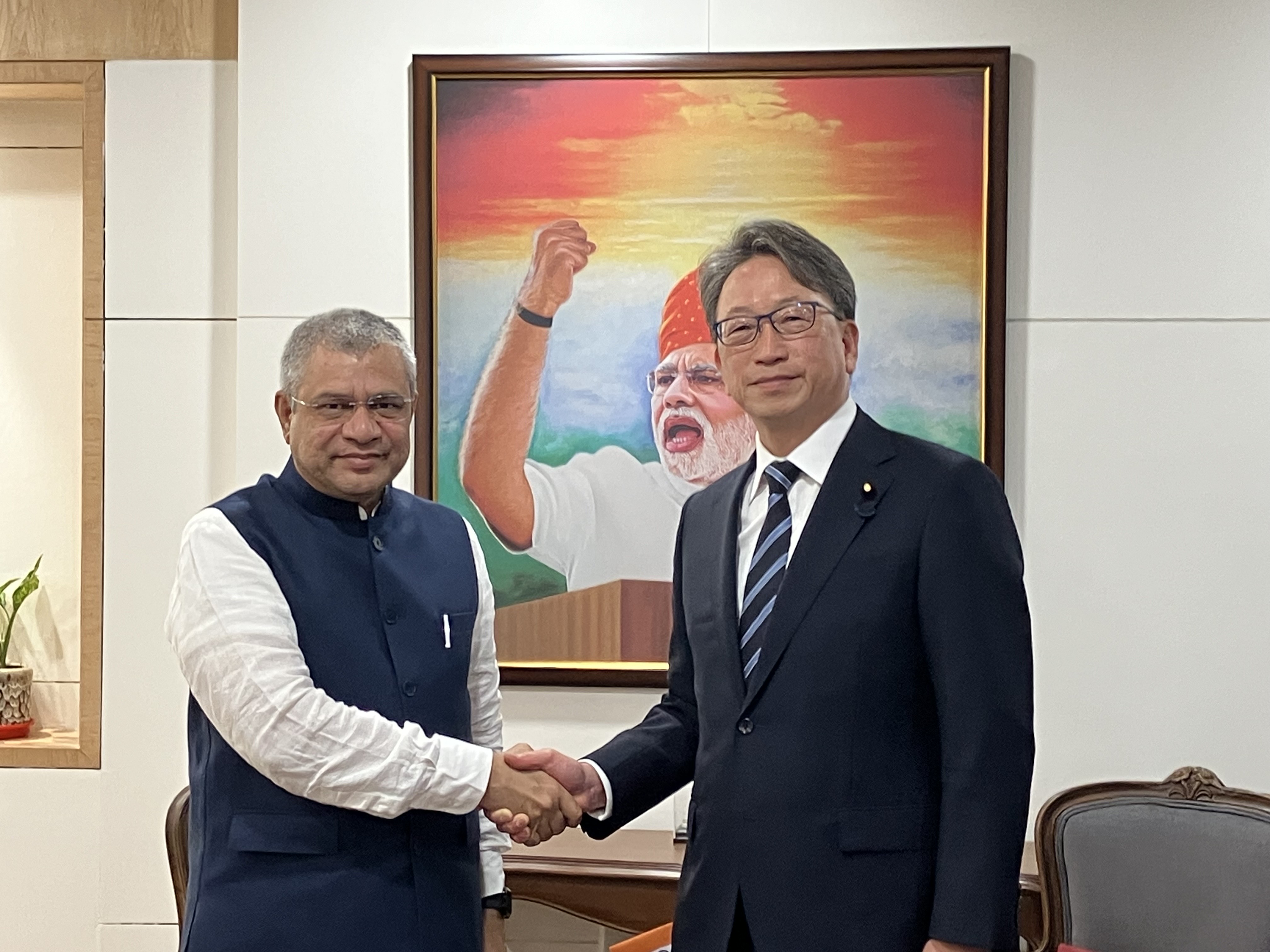 Minister Vaishnaw on the left and Minister Taira on the right shake hands during their meeting. A portrait of H.E. Mr. Narendra Modi, Prime Minister of India, is displayed behind them.