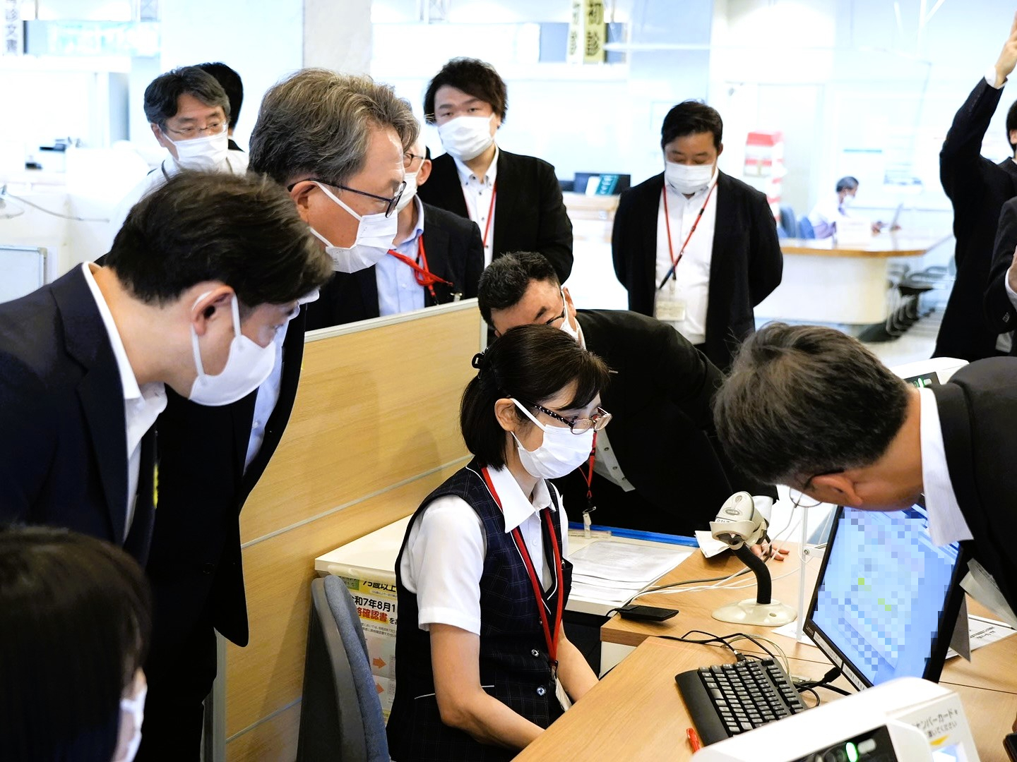 There is a monitor screen on the desk, and a person is sitting in front of the monitor. Mr. Taira, Minister for Digital Transformation, and Mr. Fukuoka, Minister of Health, Labour and Welfare, are standing behind the person and looking at the monitor.