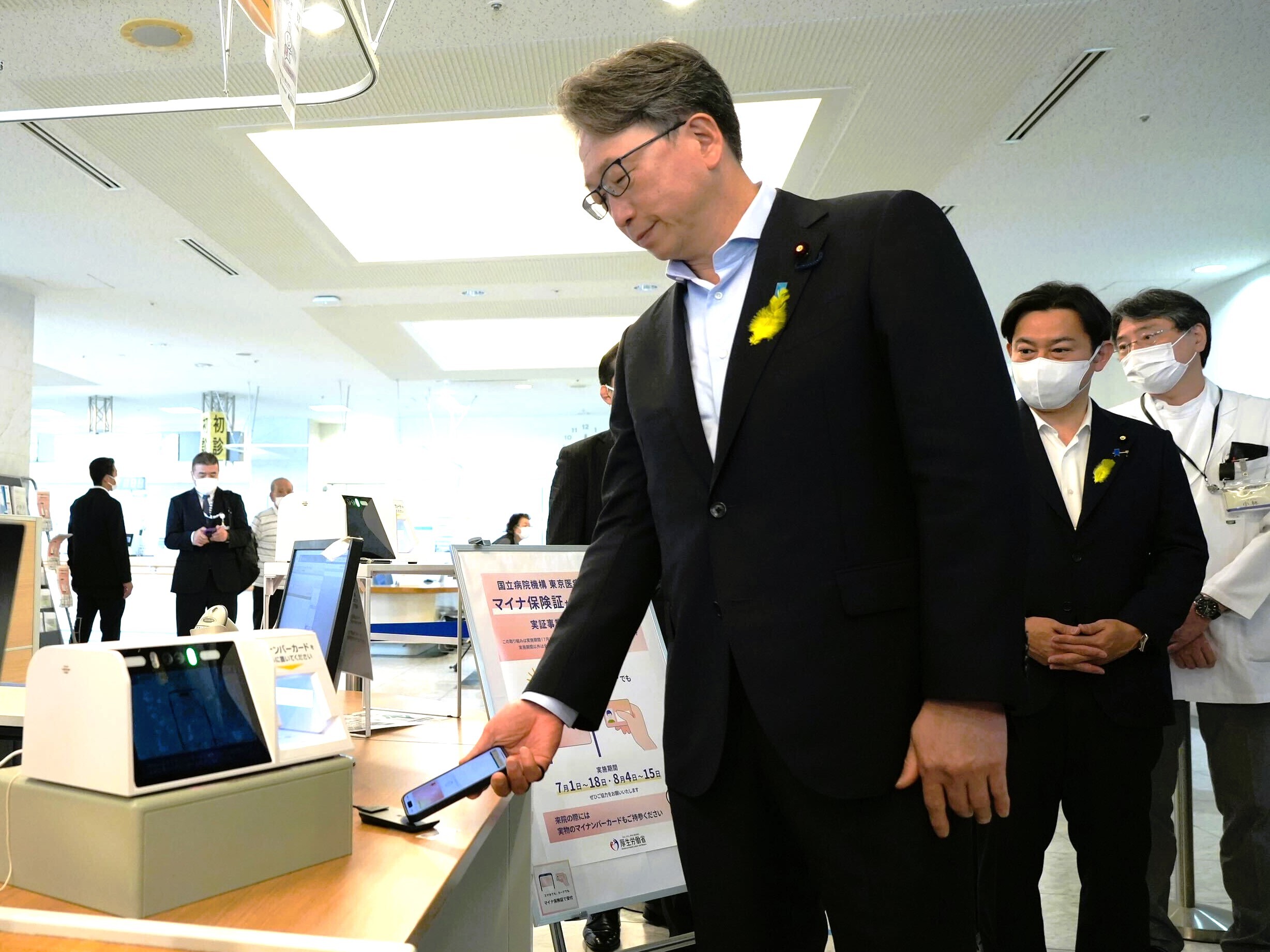 This photo shows Mr. Taira, Minister for Digital Transformation, holding his smartphone (iPhone) with his right hand over a universal card reader for smartphones. The iPhone's My Number Card is displayed on the iPhone's screen.
