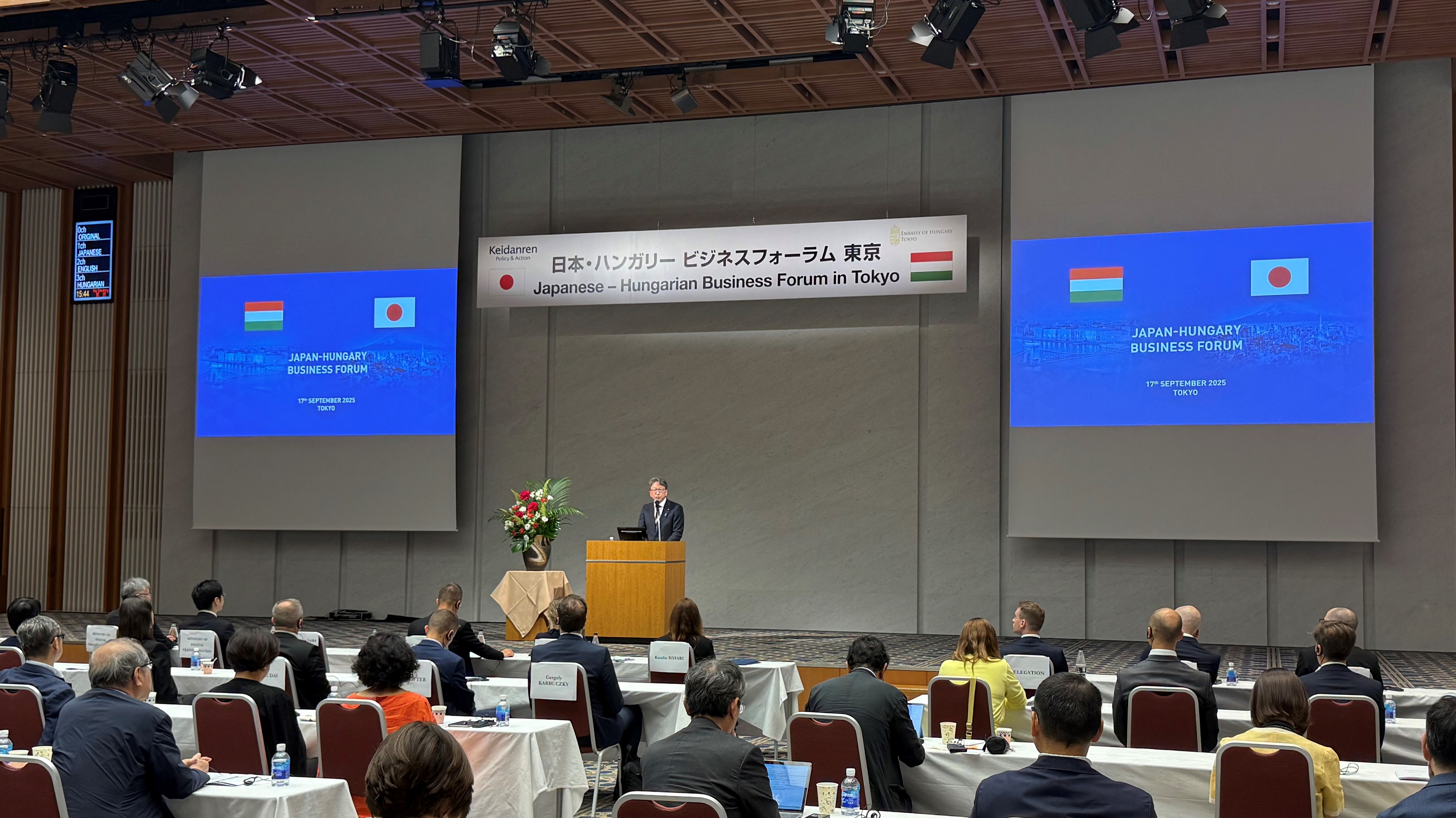 Digital Minister Taira delivering a speech at the Japanese-Hungarian Business Forum in Tokyo. In the center of the photo, Minister Taira stands on the podium addressing the audience, with the forum’s banner in the background.
