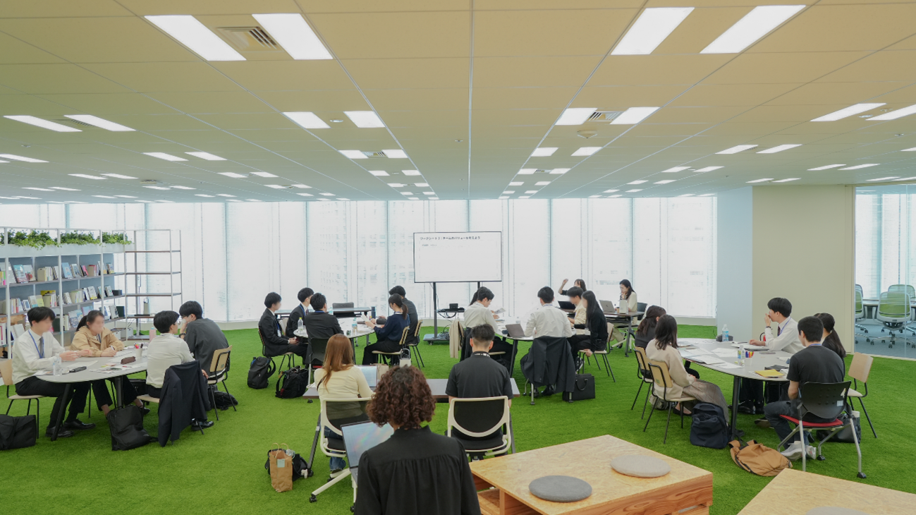 In a well-lit office space, many people are divided into several round tables to discuss and work.
In the back, there is a screen and a person who explains near it, and there are materials and laptops around.