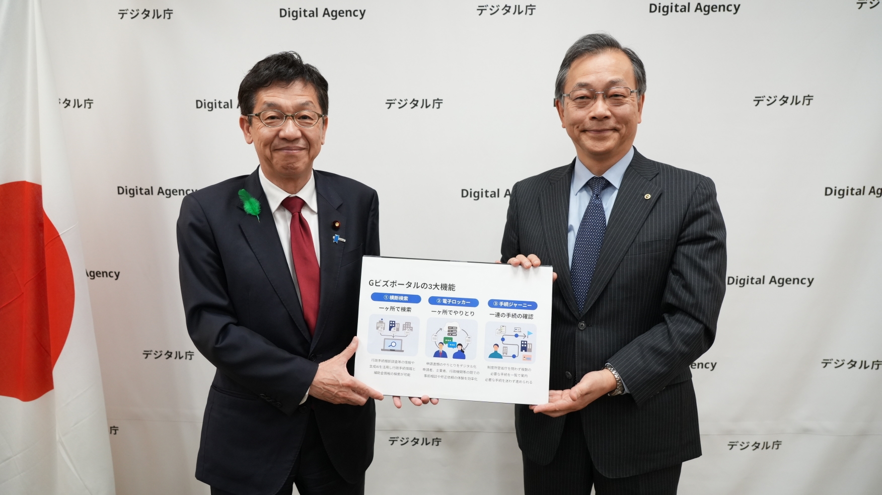 Minister for Digital Transformation Matsumoto on the left, and Executive Managing Director Hitoshi Ito of the Japan Chamber of Commerce and Industry on the right, standing side-by-side. They are jointly holding up the same white information board with diagrams with the title saying “gBizPortal”. Both men are wearing dark suits; Minister Matsumoto wears a red tie, and Executive Managing Director Ito wears a dark blue tie. They are standing in front of a white step-and-repeat backdrop covered in the Digital Agency logo, with a Japanese flag partially visible on the far left.