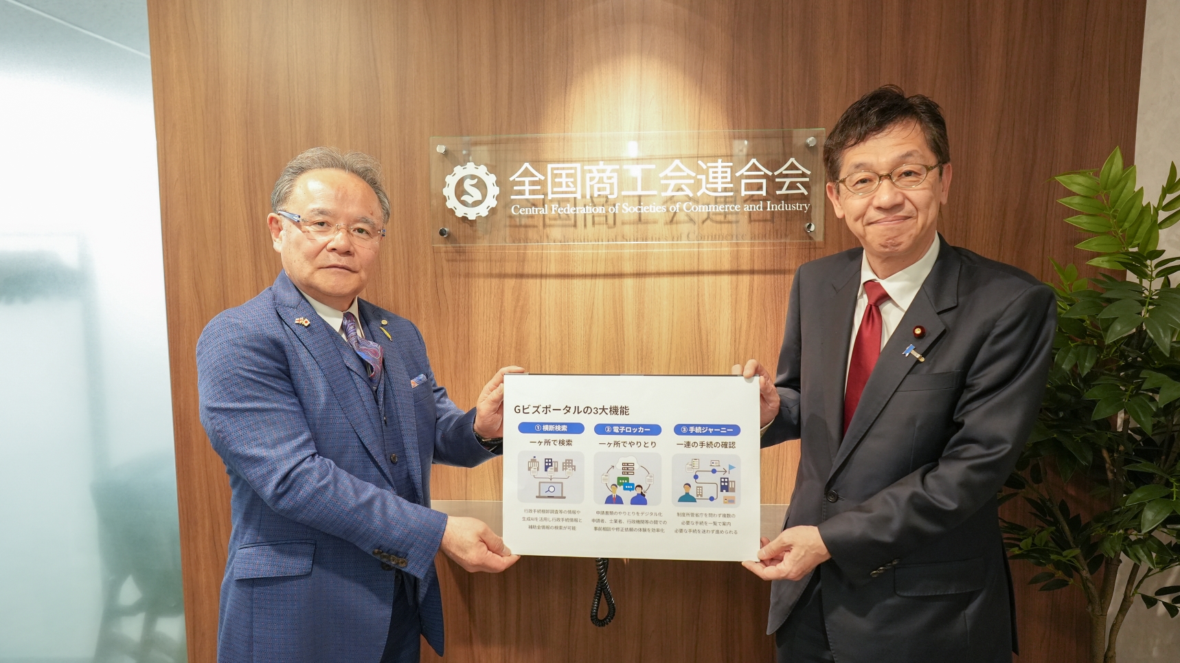 Chairman Yoshihisa Mori of the Central Federation of Societies of Commerce and Industry on the left, and Minister for Digital Transformation Matsumoto on the right, standing side-by-side indoors. They hold together a white information board displaying three diagrams with the title saying “gBizPortal”. Chairman Mori is wearing a blue suit, and Minister Matsumoto is wearing a dark suit with a red tie. Behind them is a wood-paneled wall featuring the logo and nameplate of the Central Federation of Societies of Commerce and Industry.