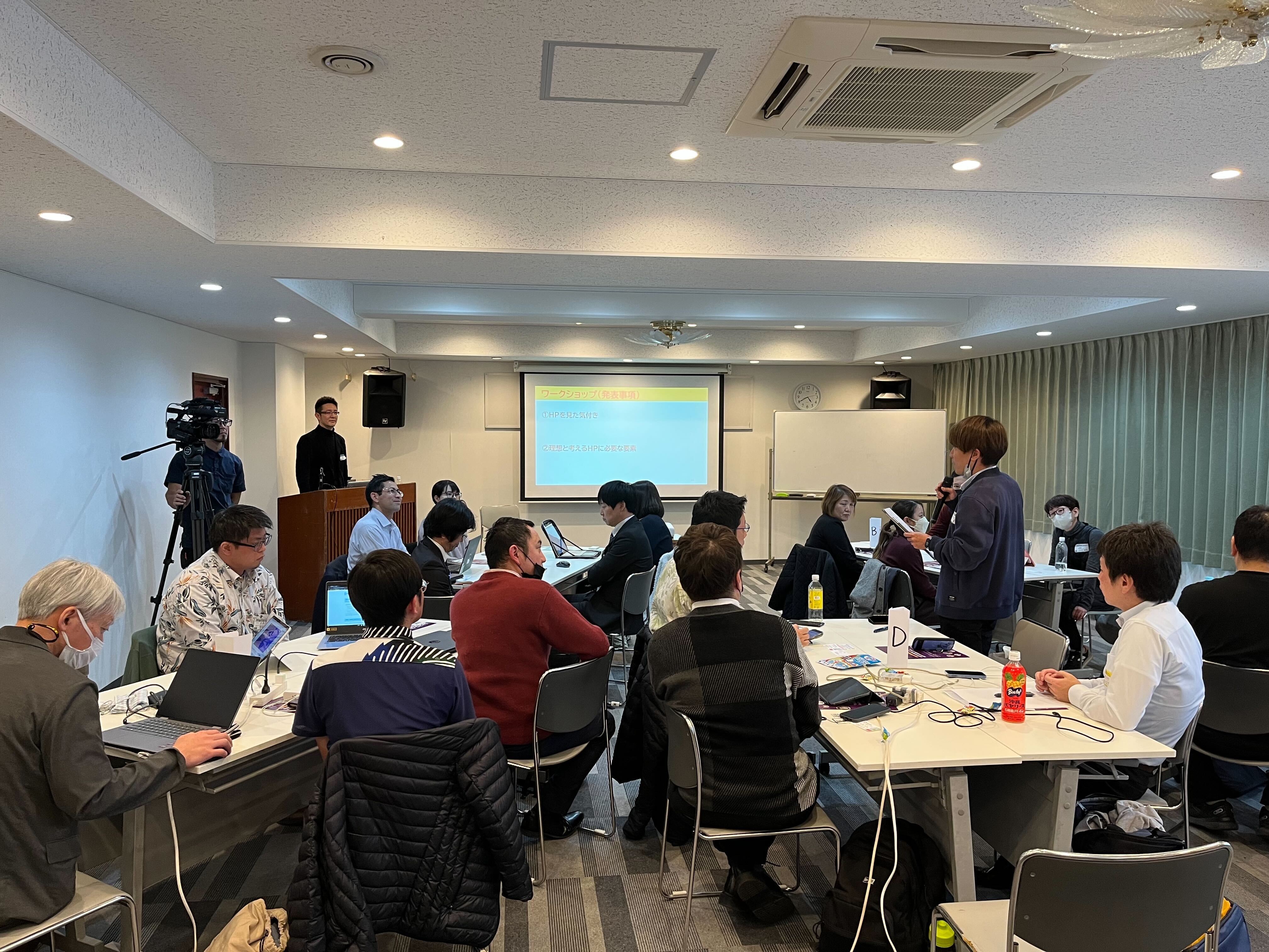 This is a photo of the co-creation PF workshop held in Naha. A presentation is projected on a screen in the back of the meeting room, and one presenter is standing with a microphone and giving a presentation to the participants. About 15 to 20 participants are divided into several groups and gathered at their desks. On the left, a photographer stands with shooting equipment and photographs the presenter. 