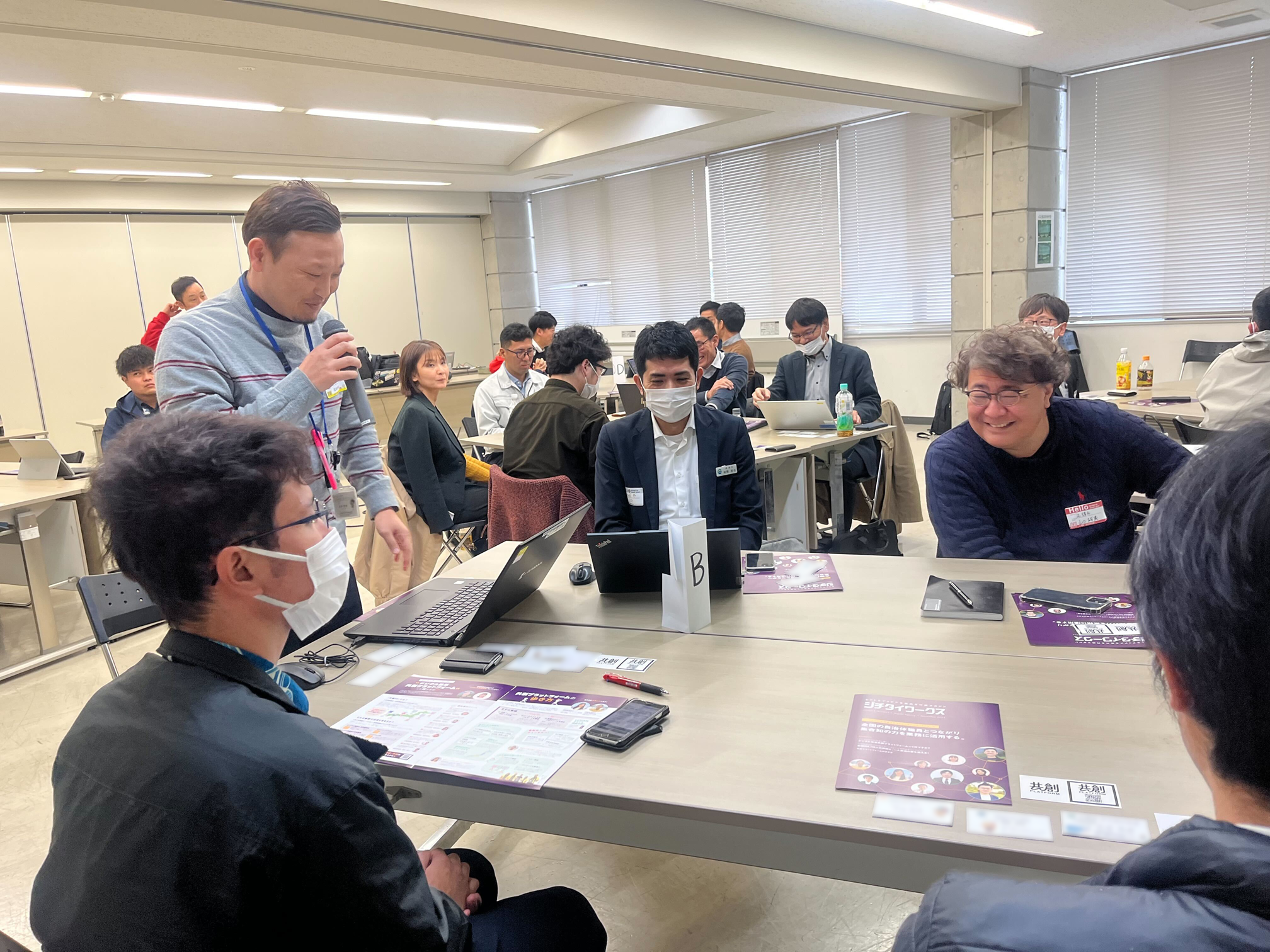 This is a photo of a co-creation PF workshop held in Nago City. One presenter is standing with a microphone and is giving a presentation while looking at the PC at hand. On the desk is a tag with a "B" written on it that represents the name of the group. The group consists of five participants including the presenter. The venue is a large conference room, and two or three other groups are gathered behind and to the left and right of each desk. 
