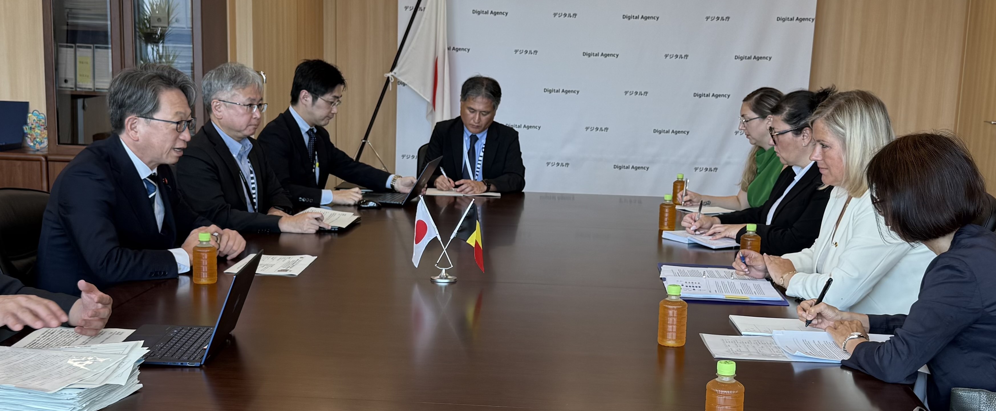 Minister for Digital Transformation Taira (left) having a meeting with Ms. Vanessa Matz (right) at his office. Mini flags of Japan and Belgium are at the center of the table, with ministers and their teams present.