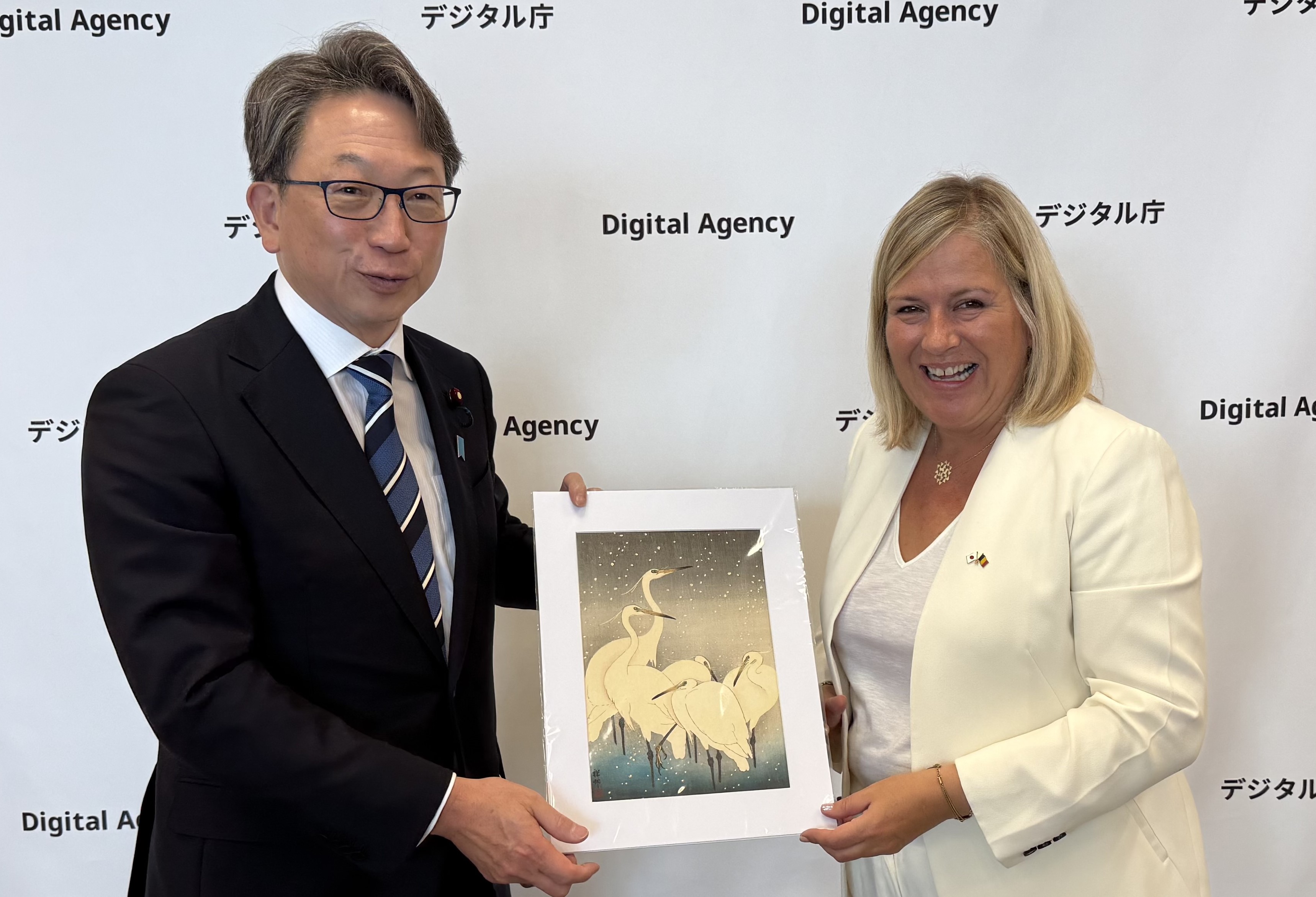 Minister for Digital Transformation Taira (left) receives a replica of Japanese “Ukiyo-e” from a Belgian museum collection, presented by Ms. Vanessa Matz, Minister of Public Modernisation, Civil Service, Public Enterprises, Digitalisation and Buildings Administration of Belgium (right). The background shows the Digital Agency logo panel. Both ministers smile while holding a replica of Koson Ohara’s Ukiyo-e (painting) of multiple cranes standing in the snow.