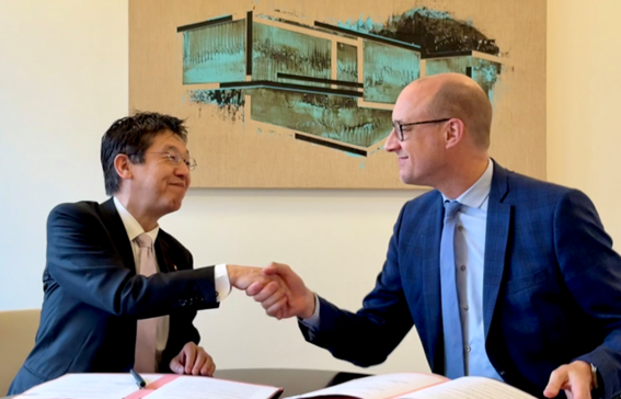 Minister for Digital Transformation Matsumoto (left) and Deputy Prime Minister Vincent Van Peteghem (right) sitting in chairs and shaking hands. Two copies of the Memorandum of Cooperation and a pen are placed on the desk.