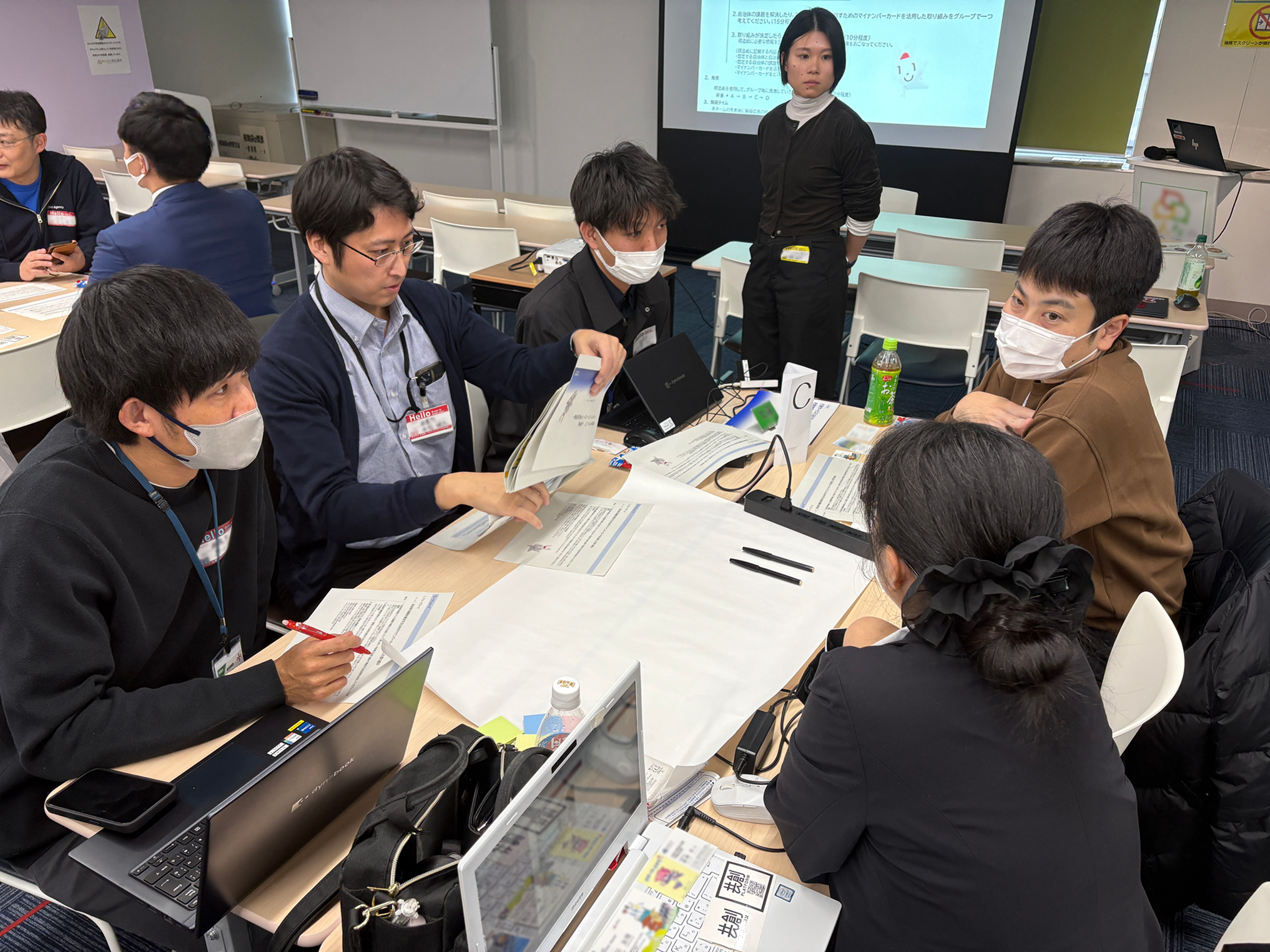 A photo of the workshop, showing five people talking around a desk with a sheet of imitation paper spread out in the middle. The five people are sitting looking at a document.