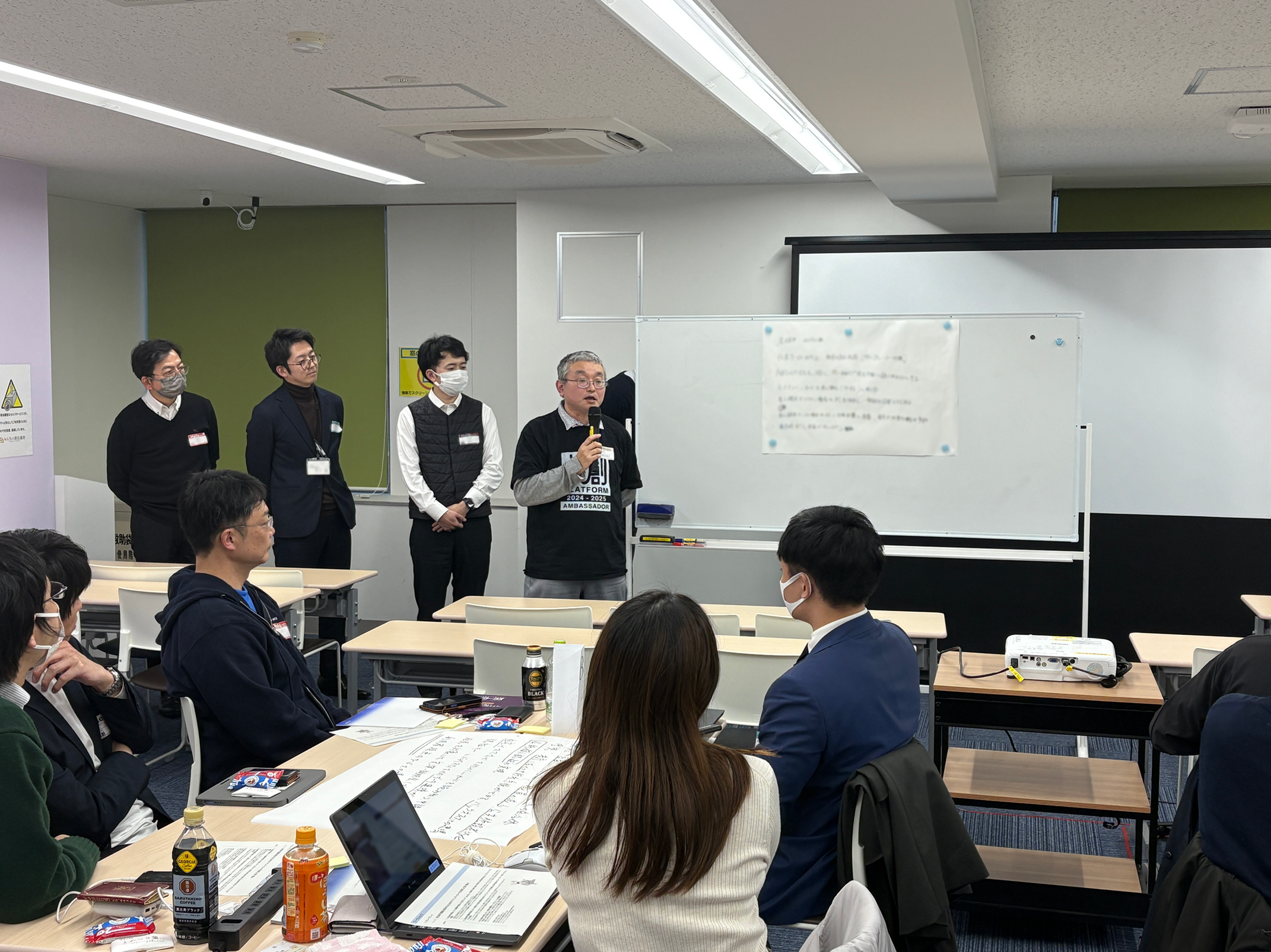 In this photo, four people are giving a presentation at a workshop. One of them is holding a microphone and giving a presentation. In front of the presenter, five people are sitting around a desk and listening to the presentation.