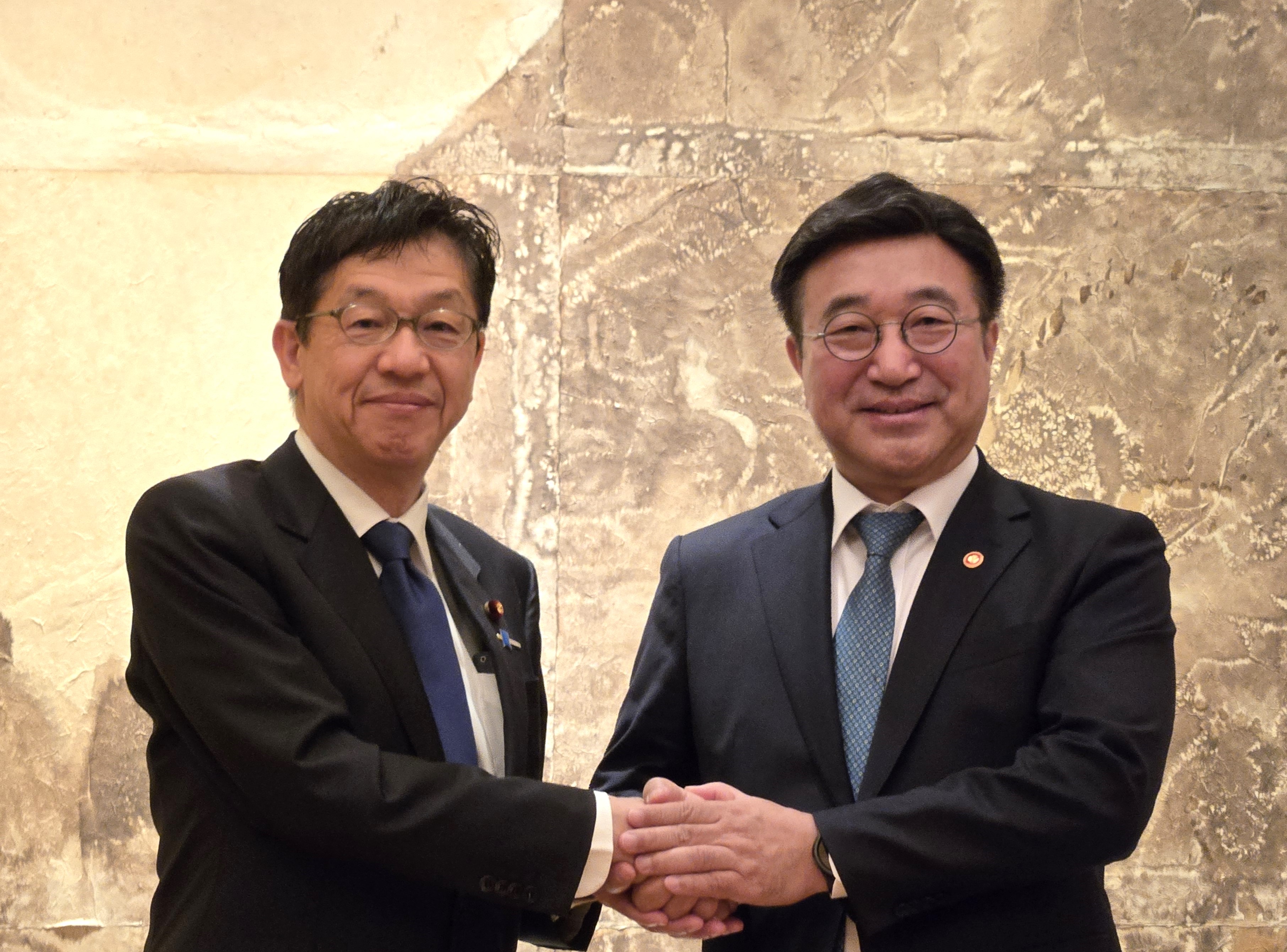 Minister for Digital Transformation Matsumoto (left) and Minister of the Interior and Safety, Republic of Korea Yun Hojung (right), both wearing eyeglasses, dark blue suits, white shirts, and light blue ties, smiling and shaking hands in front of a marble wall.