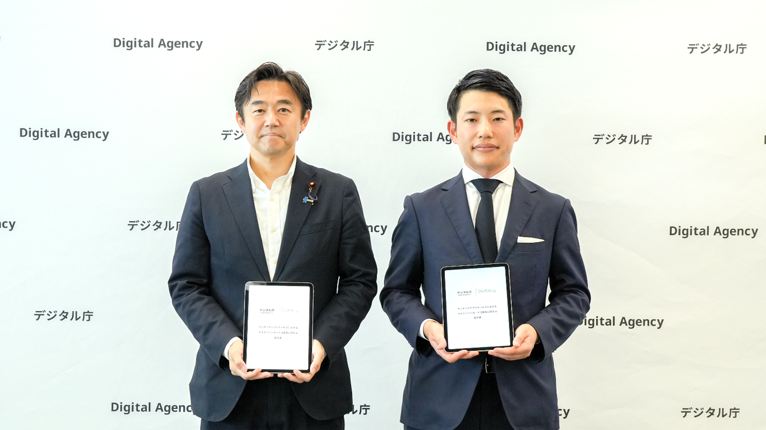 Mr. Hosaka, State Minister for Digital Transformation, is standing on the left, and Iizuka, the representative director of the Association of Matching Apps for Love and Marriage, is standing on the right, with a white back panel with multiple Digital Agency logos on it. They are holding tablets with their corporate logos and the words 
