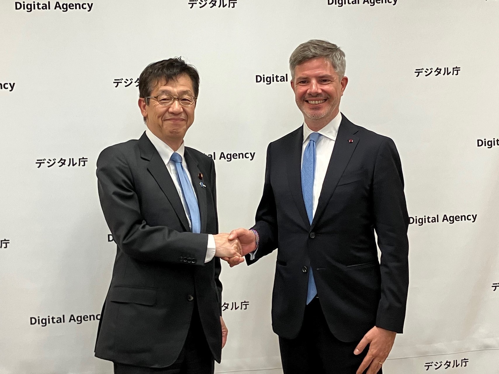 Two men in business suits stand side by side, facing the camera, and shaking hands. Both are smiling, indicating a formal meeting. On the left is Minister for Digital Transformation Matsumoto, wearing glasses, a dark suit, a white shirt, and a light blue tie. His expression is calm and composed, with a polite smile. On the right is Mr. Antoine Evrard, Ambassador of Belgium to Japan, also dressed in a dark suit, a white shirt, and a light blue tie. He is smiling warmly. They are engaged in a handshake. The background is a plain white wall patterned with the “Digital Agency” logo in both English and Japanese.