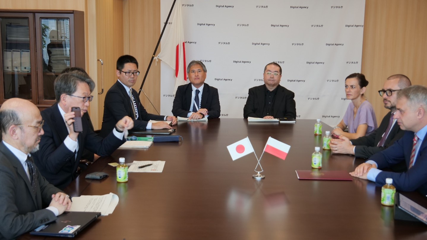 Meeting photo: Minister Taira (on the left) having a meeting with Deputy Prime Minister and Minister for Digital Affairs Gawkowski (on the right) across the table. He explains the My Number Card function on smartphones while holding smartphone and the card. The national flags of Japan and Poland are in the center.