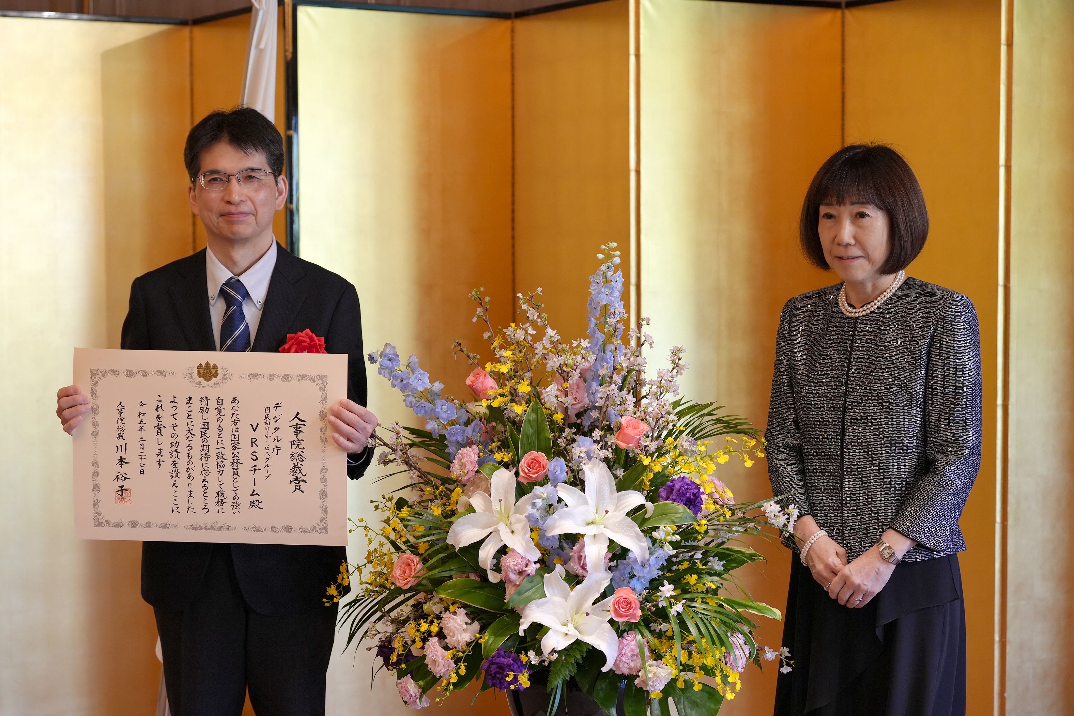From left to right, the Digital Commissioner and President Yuko Kawamoto (National Personnel Authority) stand side by side. The commissioner spreads the certificate of commendation in front of his chest and holds it with both hands.