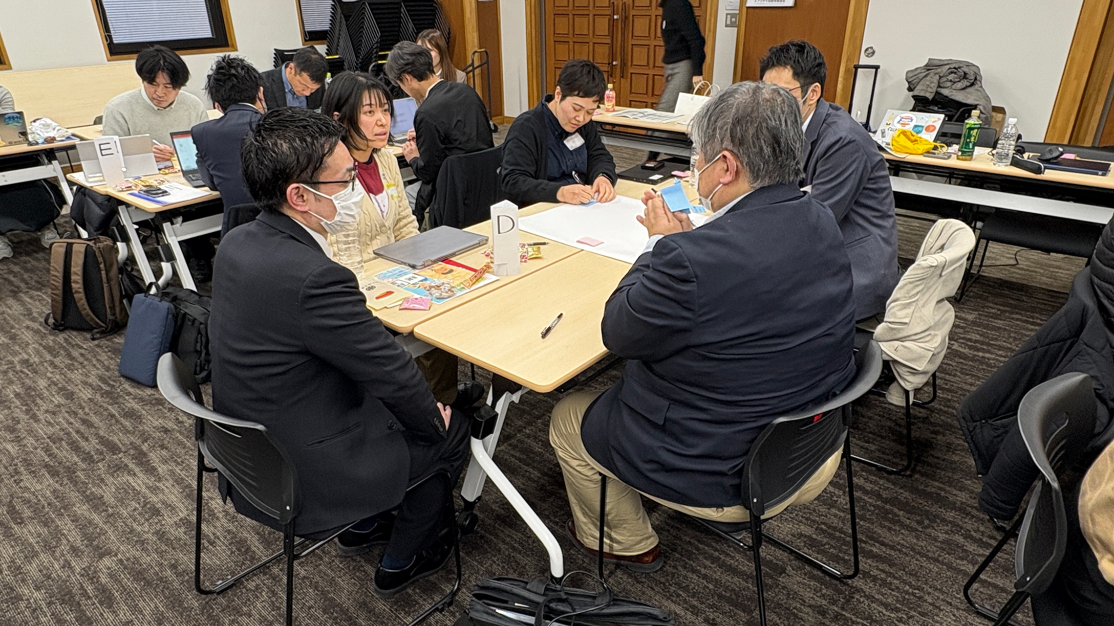 A photo of a workshop. Five people are talking around a desk. A piece of imitation paper is spread out in the middle of the desk, and one person is taking notes on the paper. A tag with a "D" written on it is on the desk.