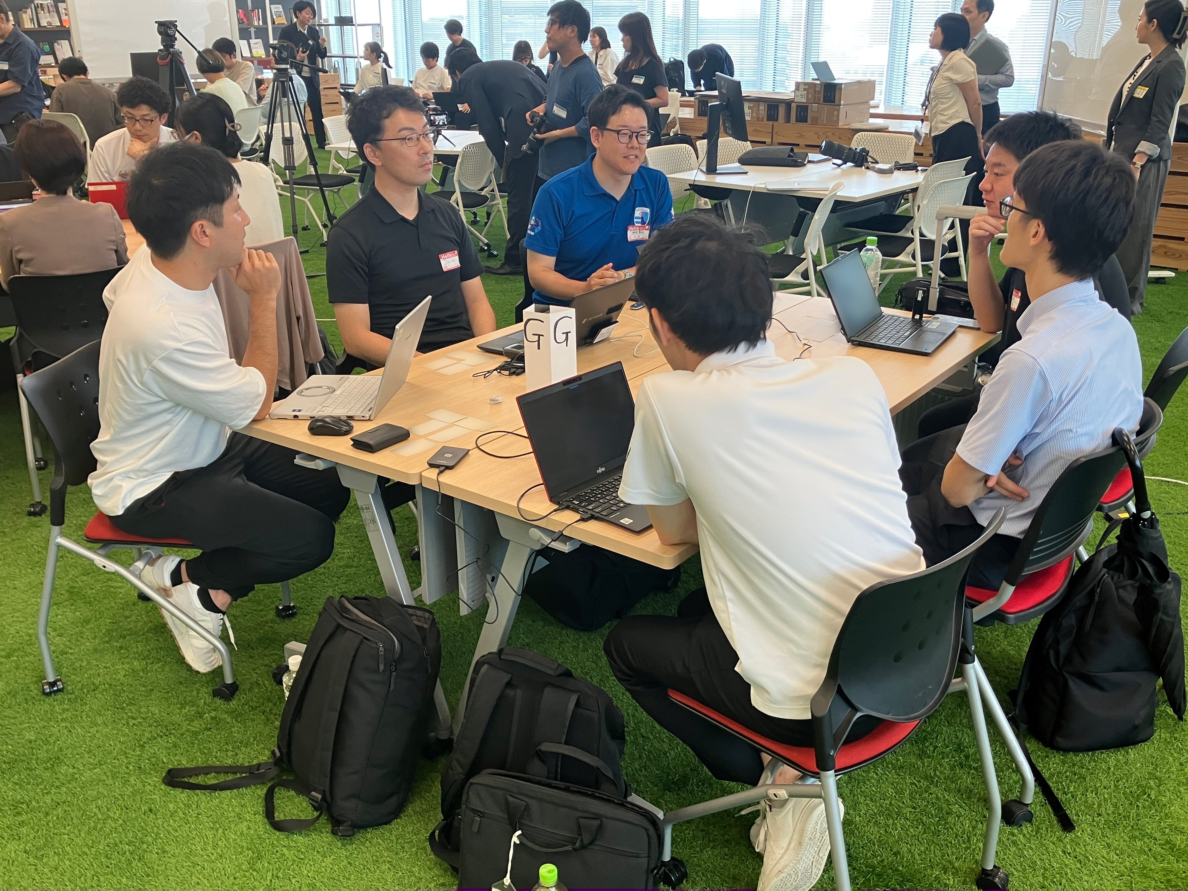The photo shows six participants discussing the use of AI in the work of local governments. On the desk, there is a tag with a "G" written on it representing the name of the group. Two or three other groups are gathered behind and to the left and right of each desk.