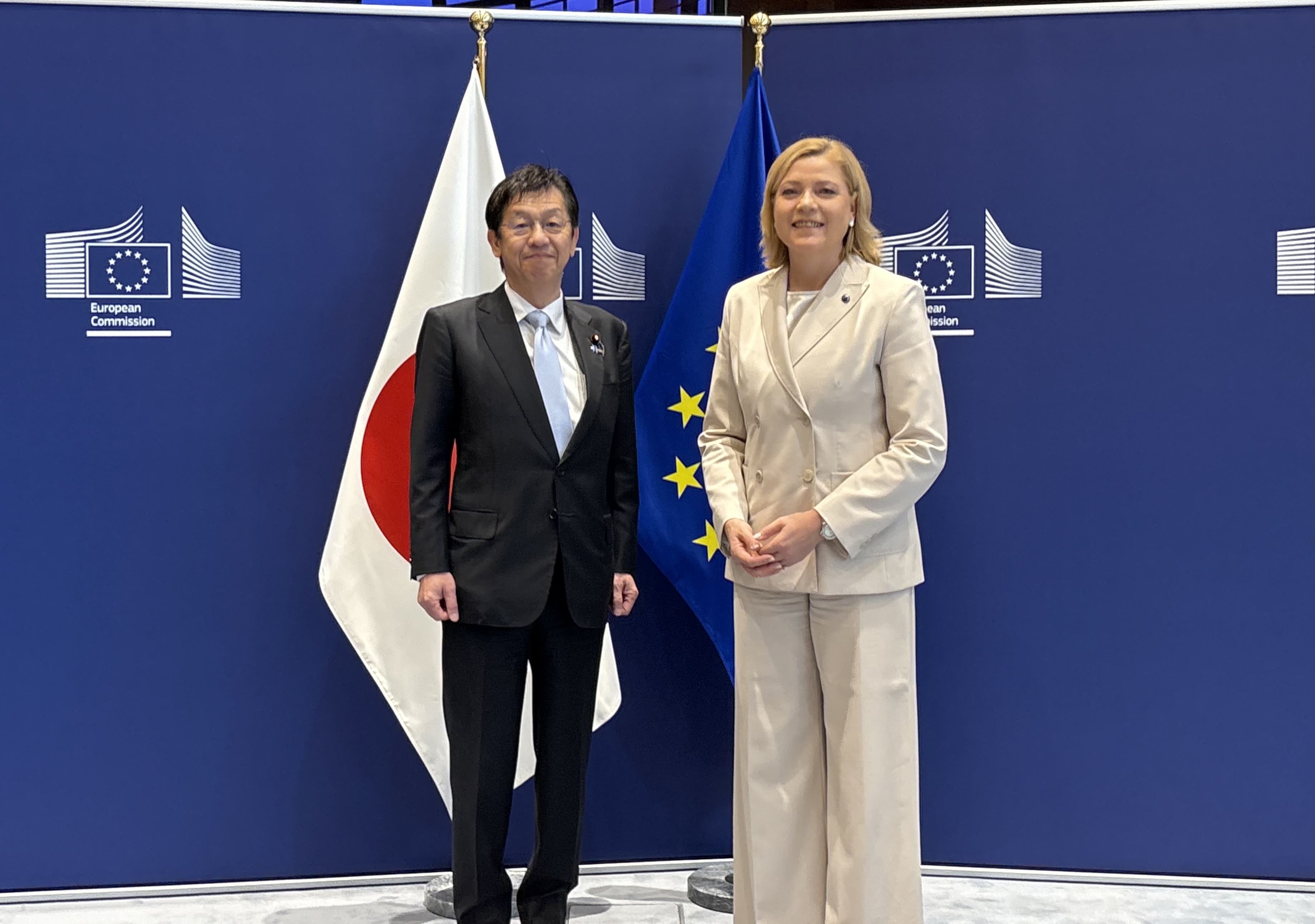 Photo of Minister Matsumoto (left) and Executive Vice-President of the European Commission Virkkunen (right) standing side-by-side, with the Japanese and EU flags in the background.