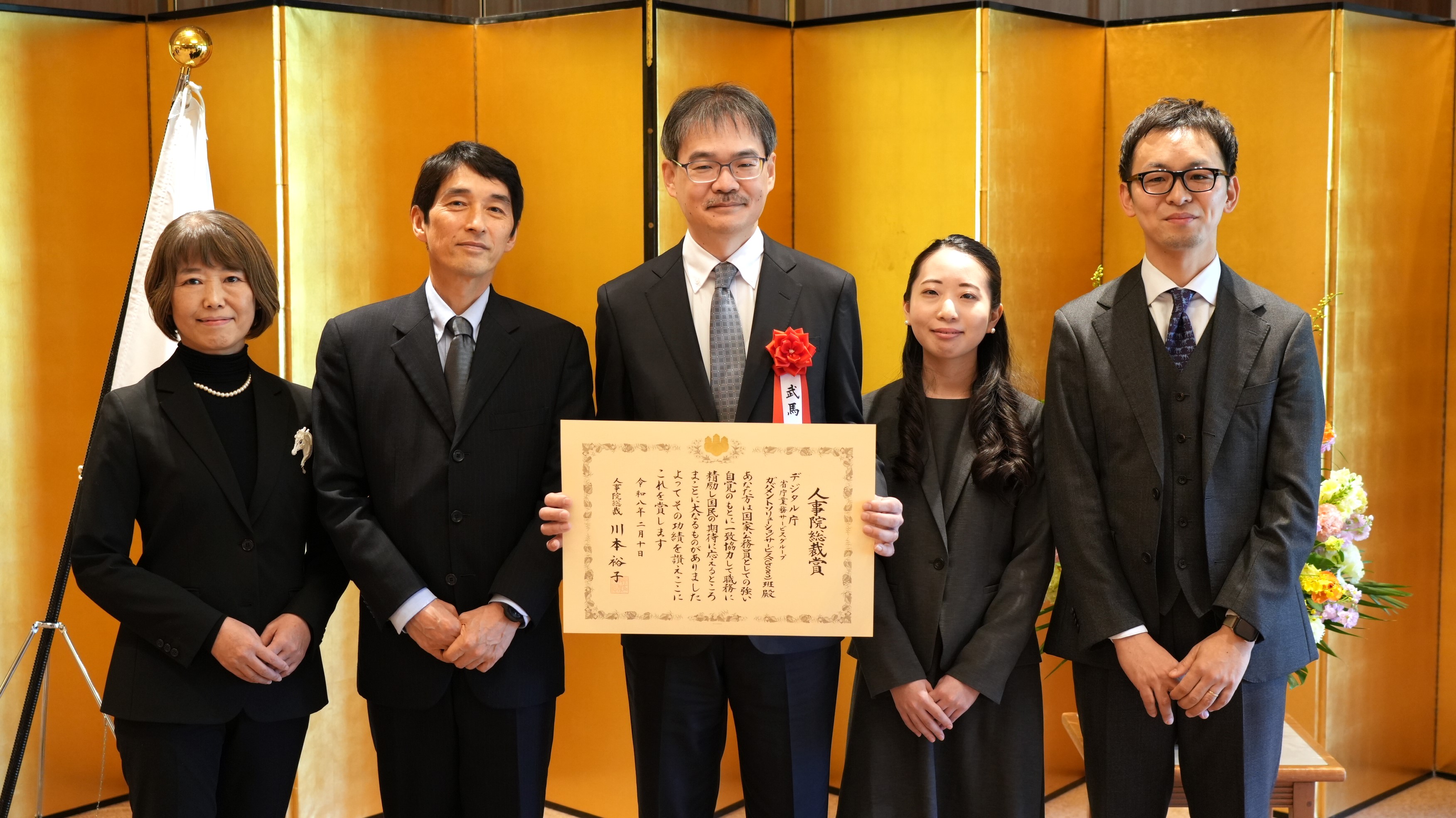 Group photo of five members of the Government Solution Services team at the National Personnel Authority President’s Award Ceremony, standing before a gold folding screen and smiling. The man in the center holding the award certificate.