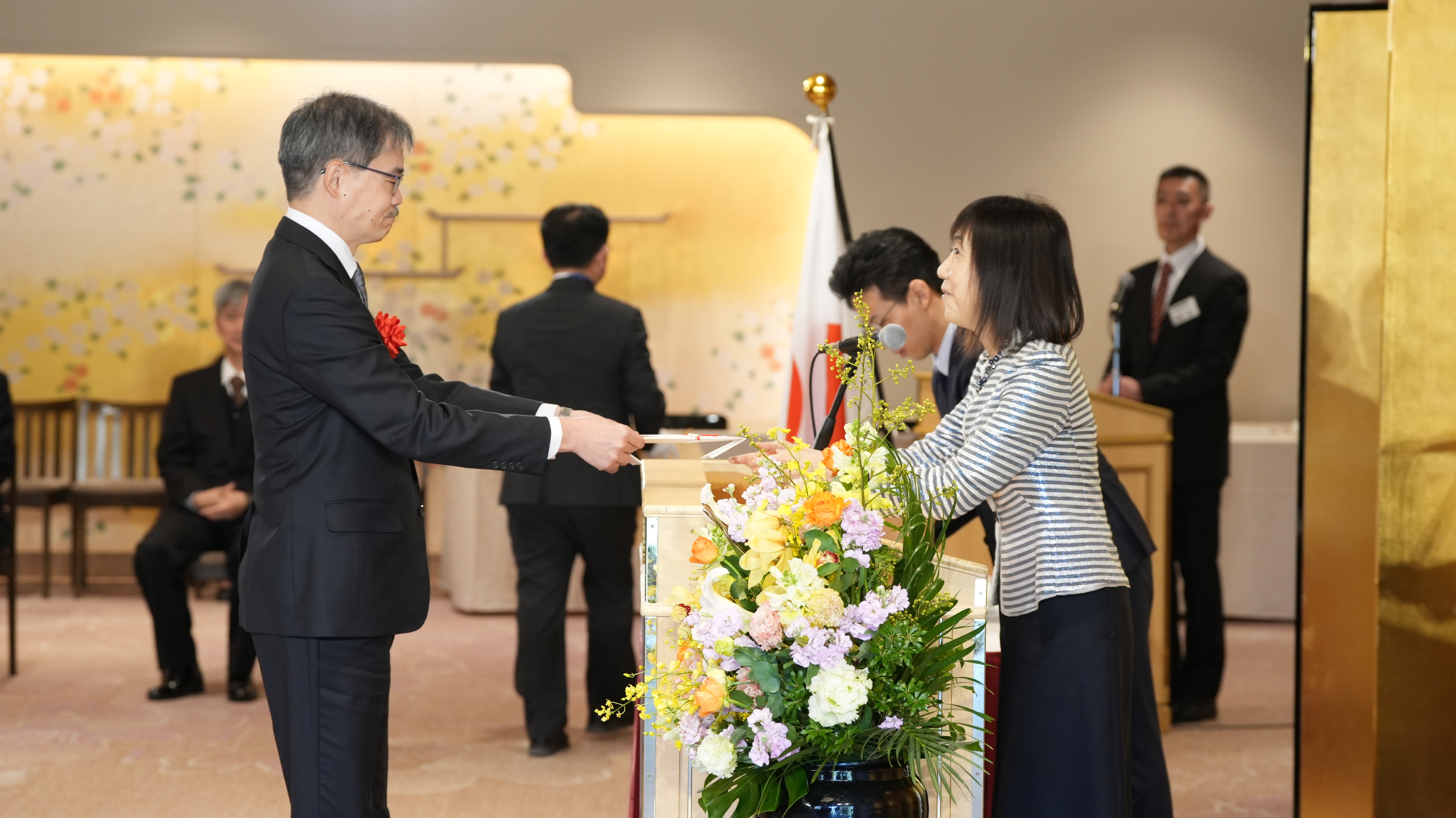 Scene from the award ceremony. A representative (woman) from the National Personnel Authority on the right presenting the certificate to the GSS team representative. The floral arrangements in purple, orange, yellow, and white in the foreground.